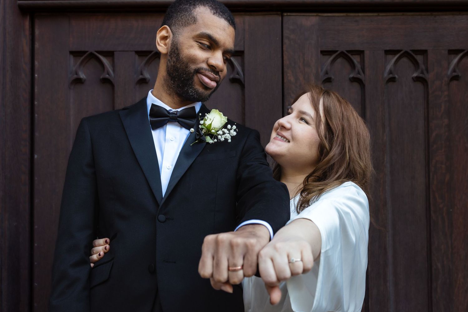 Newlywed couple smiling and showing wedding rings outside wooden church doors.
