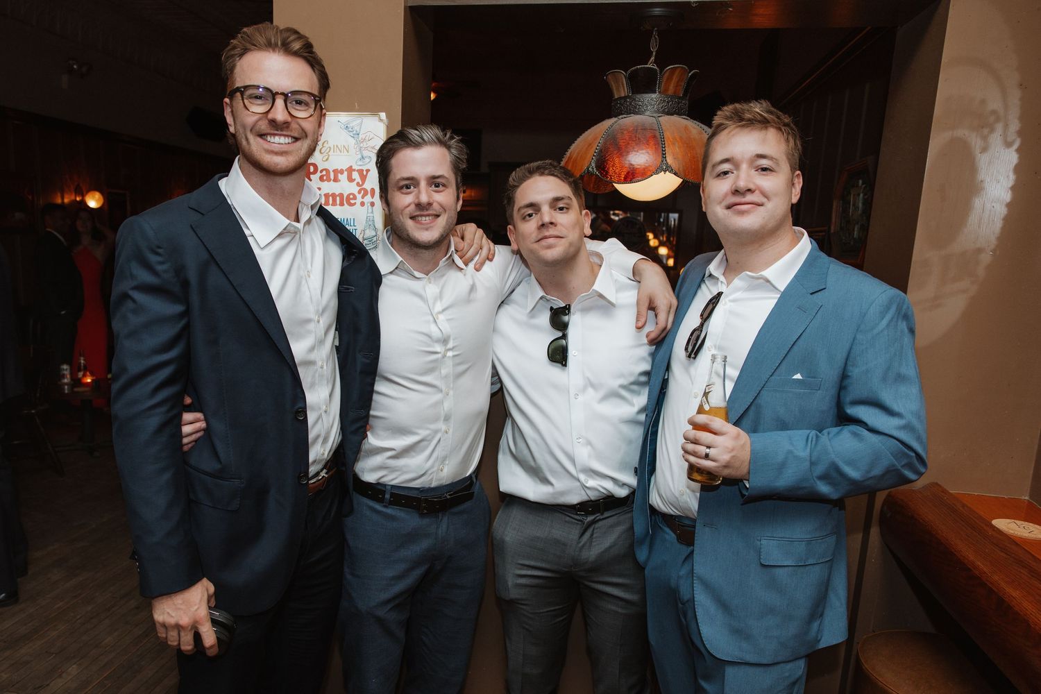 Four well-dressed men posing together and smiling at an indoor wedding reception celebration.