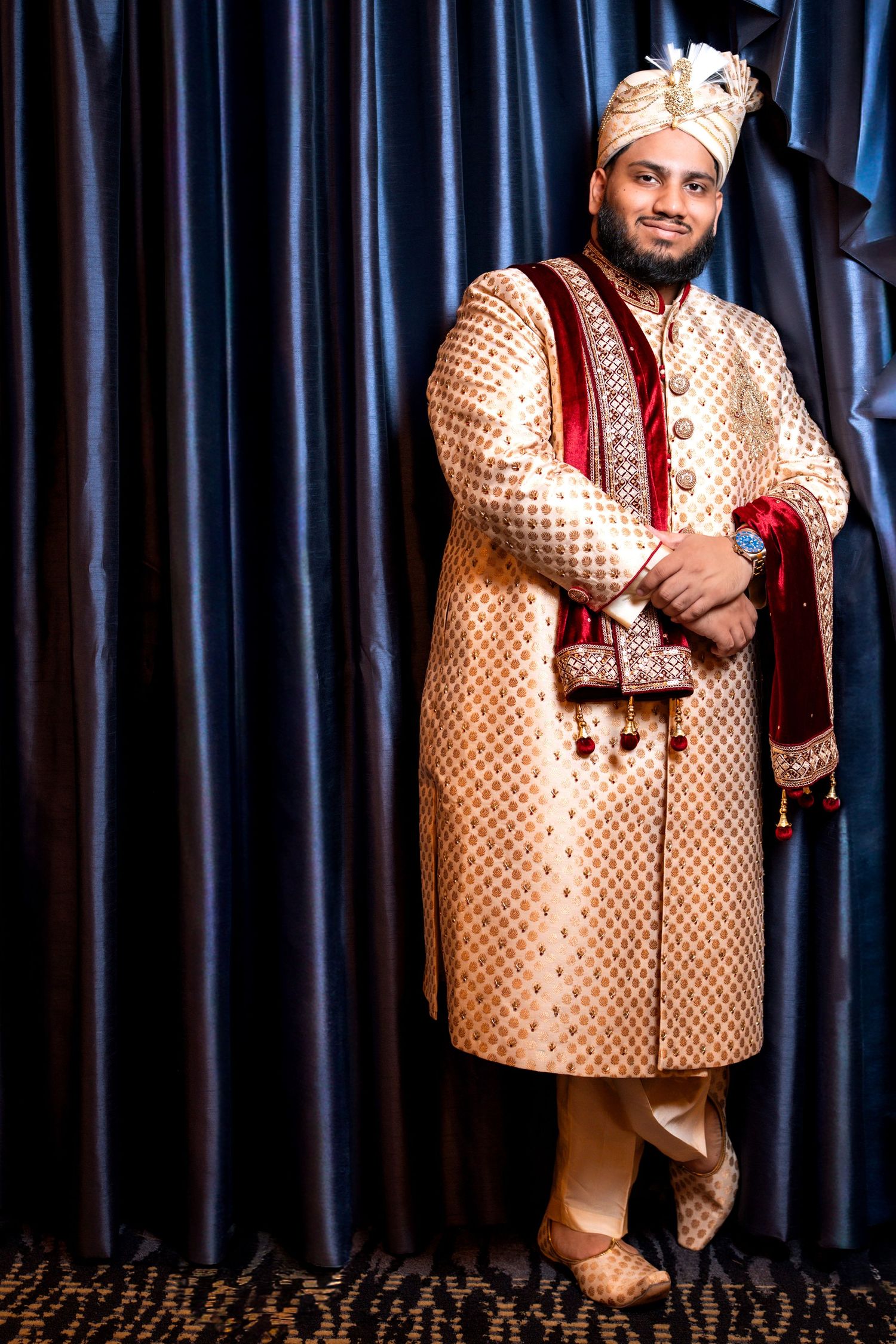 Indian groom in traditional gold sherwani and turban standing confidently against blue curtain backdrop.