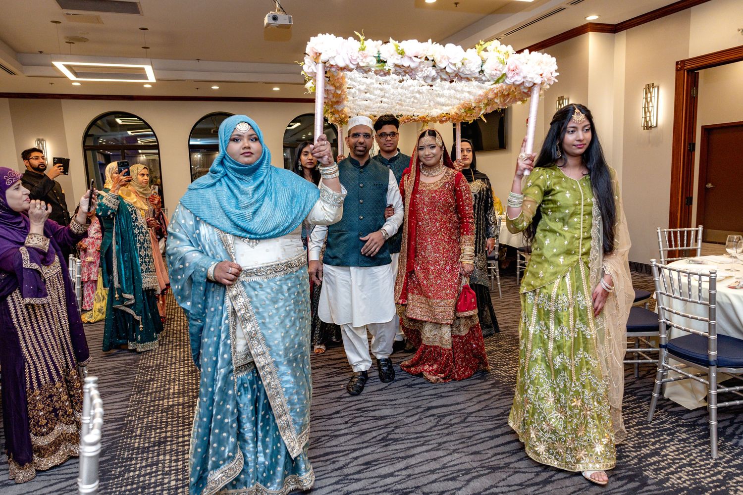 South Asian wedding procession with bride under floral canopy surrounded by family in traditional dress.