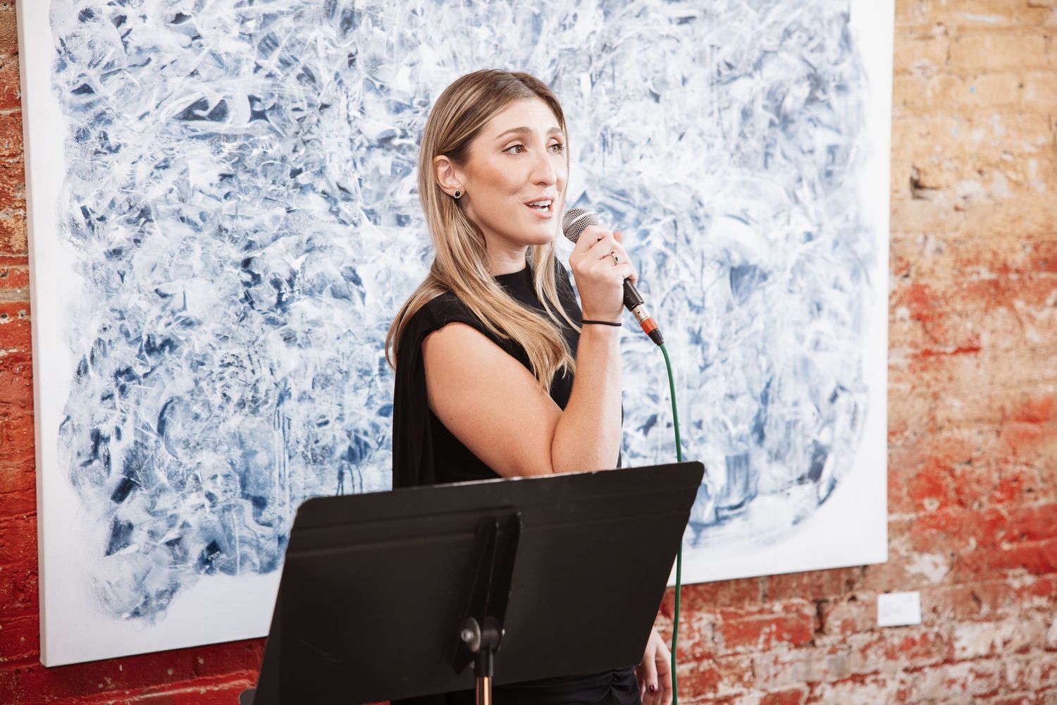 A blonde woman in a black dress speaks into a microphone at a podium in an art gallery.