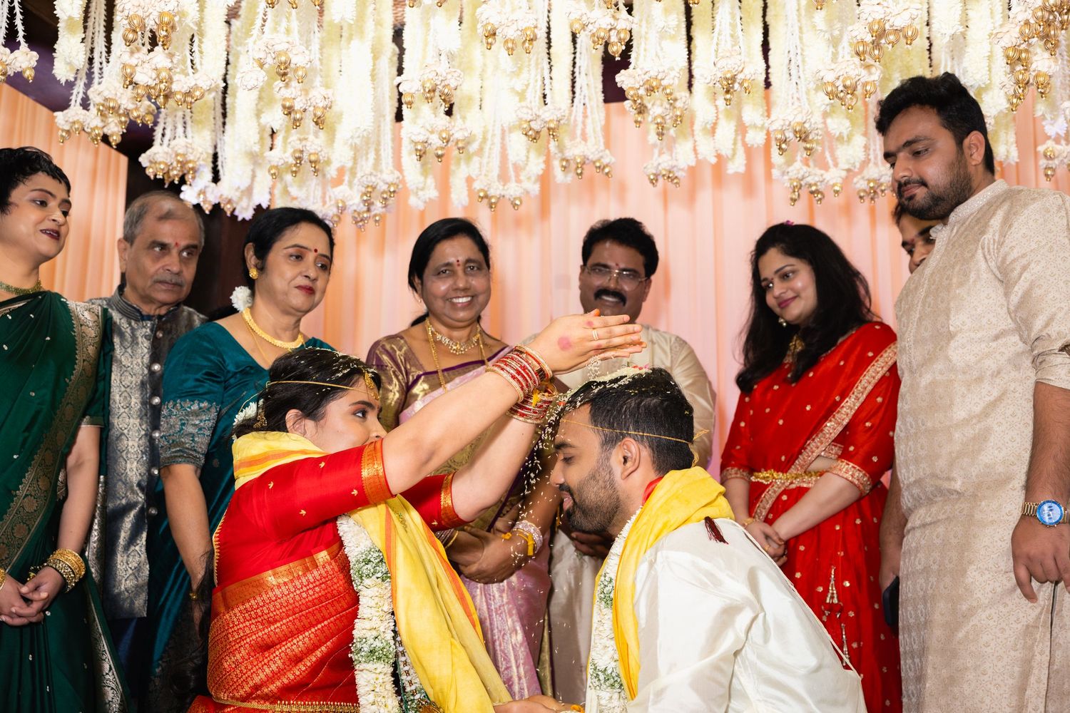 Family surrounds Indian bride and groom during traditional Hindu wedding blessing ceremony under floral decor.