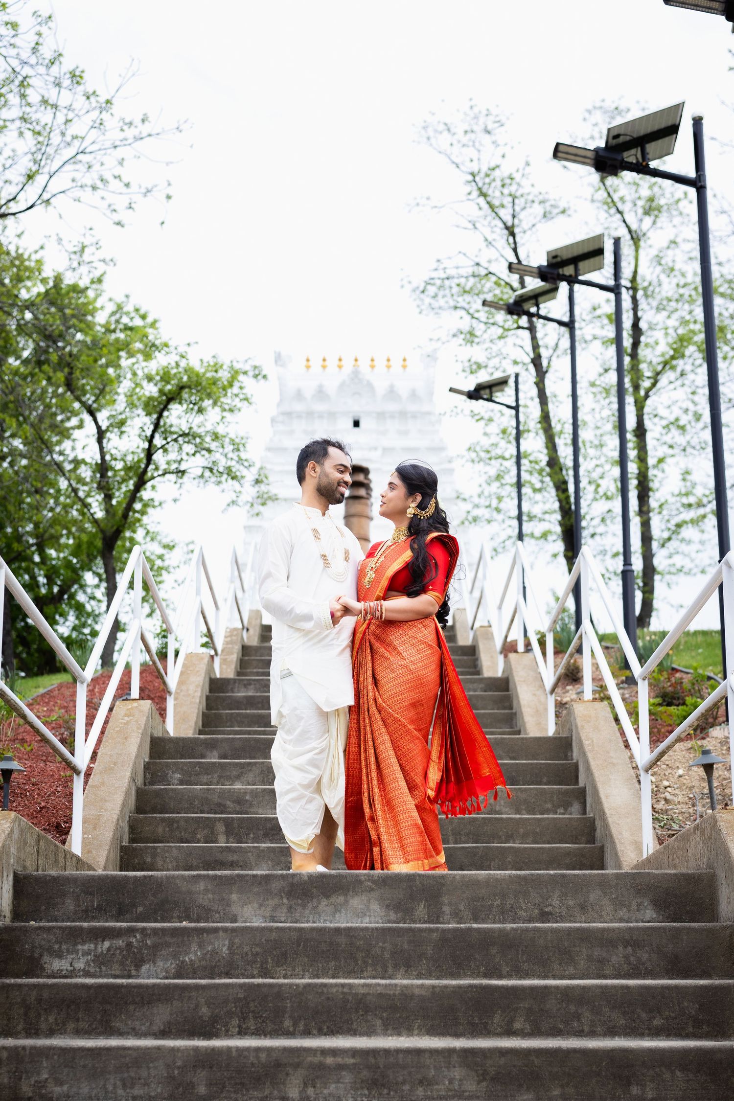 Indian wedding couple in traditional attire stand facing each other on steps with white temple backdrop.