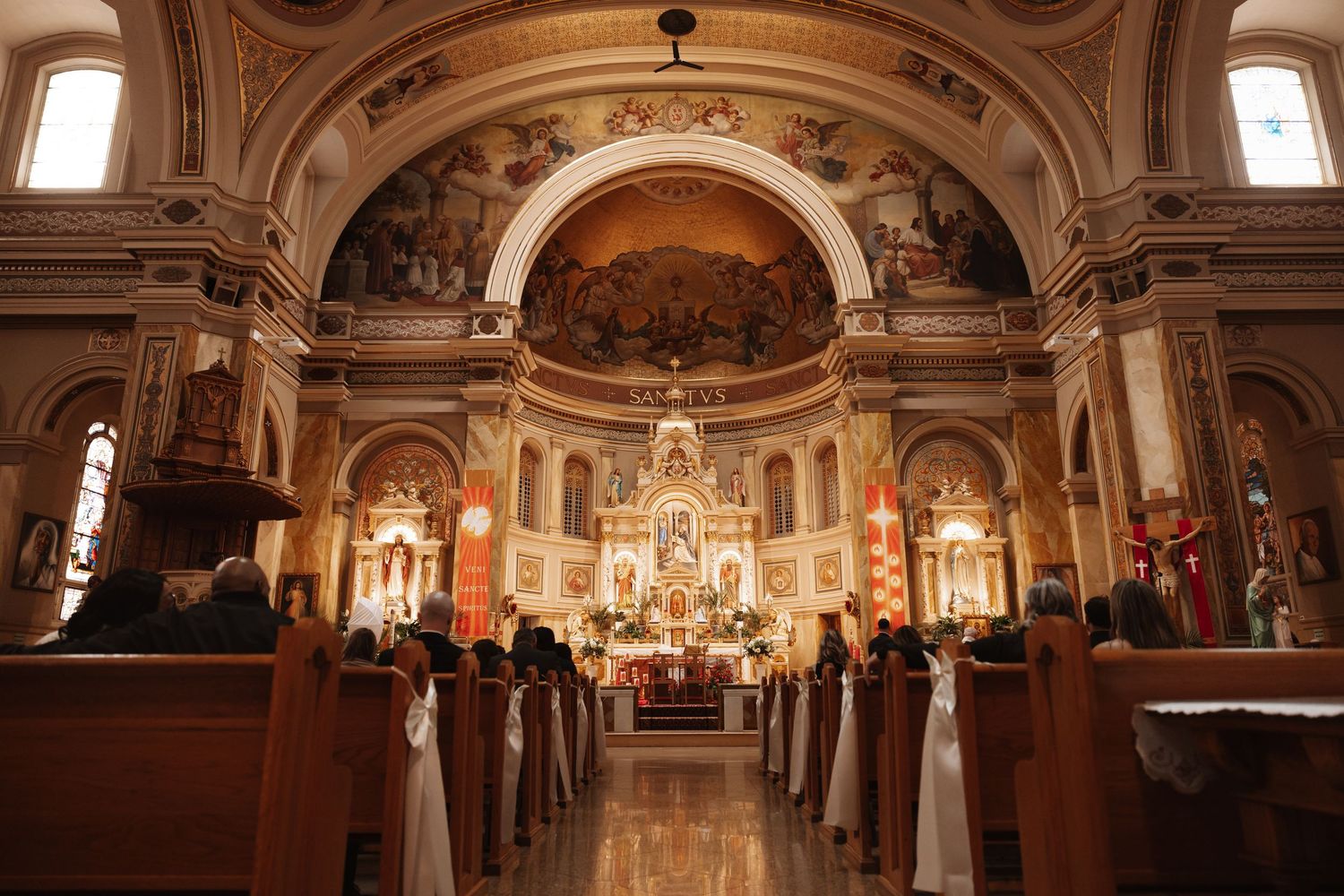 Ornate Catholic church interior filled with wedding guests seated in wooden pews beneath stunning frescoed arches.