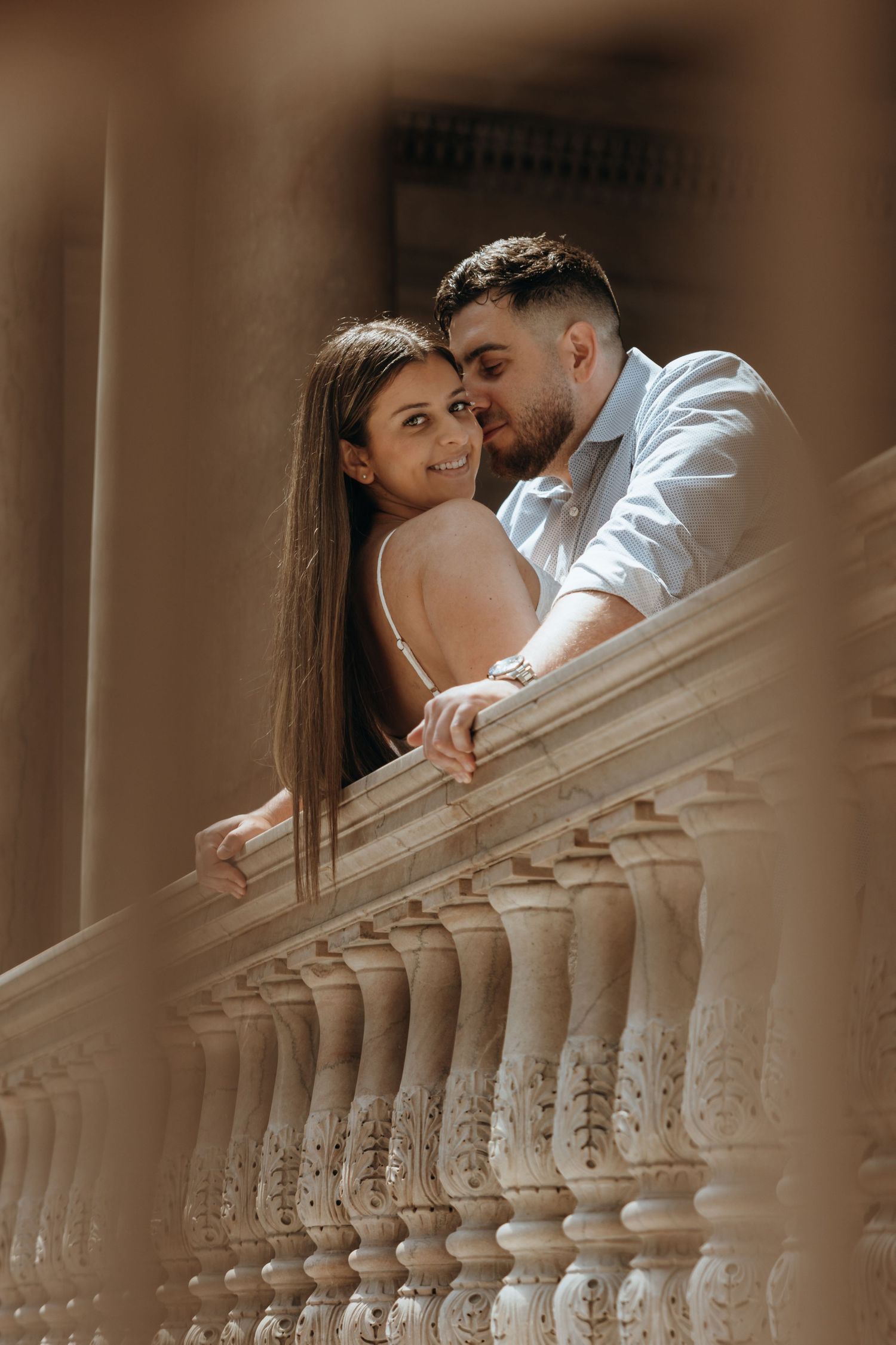 Couple sharing a romantic moment on a classical stone balustrade balcony, woman in backless dress, man in blue shirt.