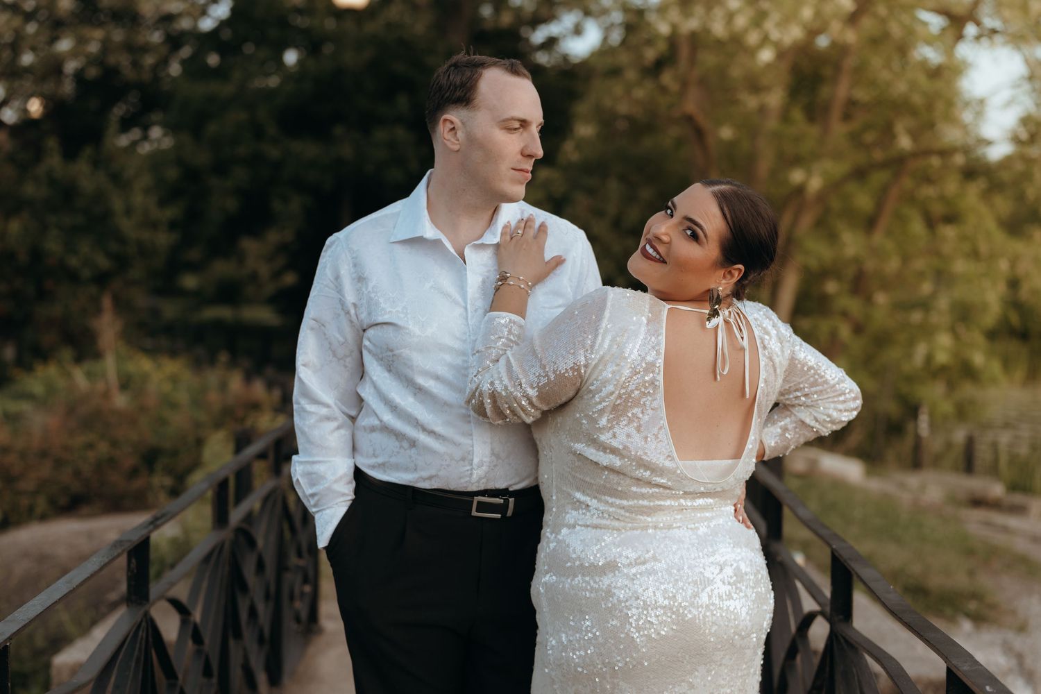 Couple sharing a joyful outdoor moment on a bridge, bride in sequined gown at golden hour.