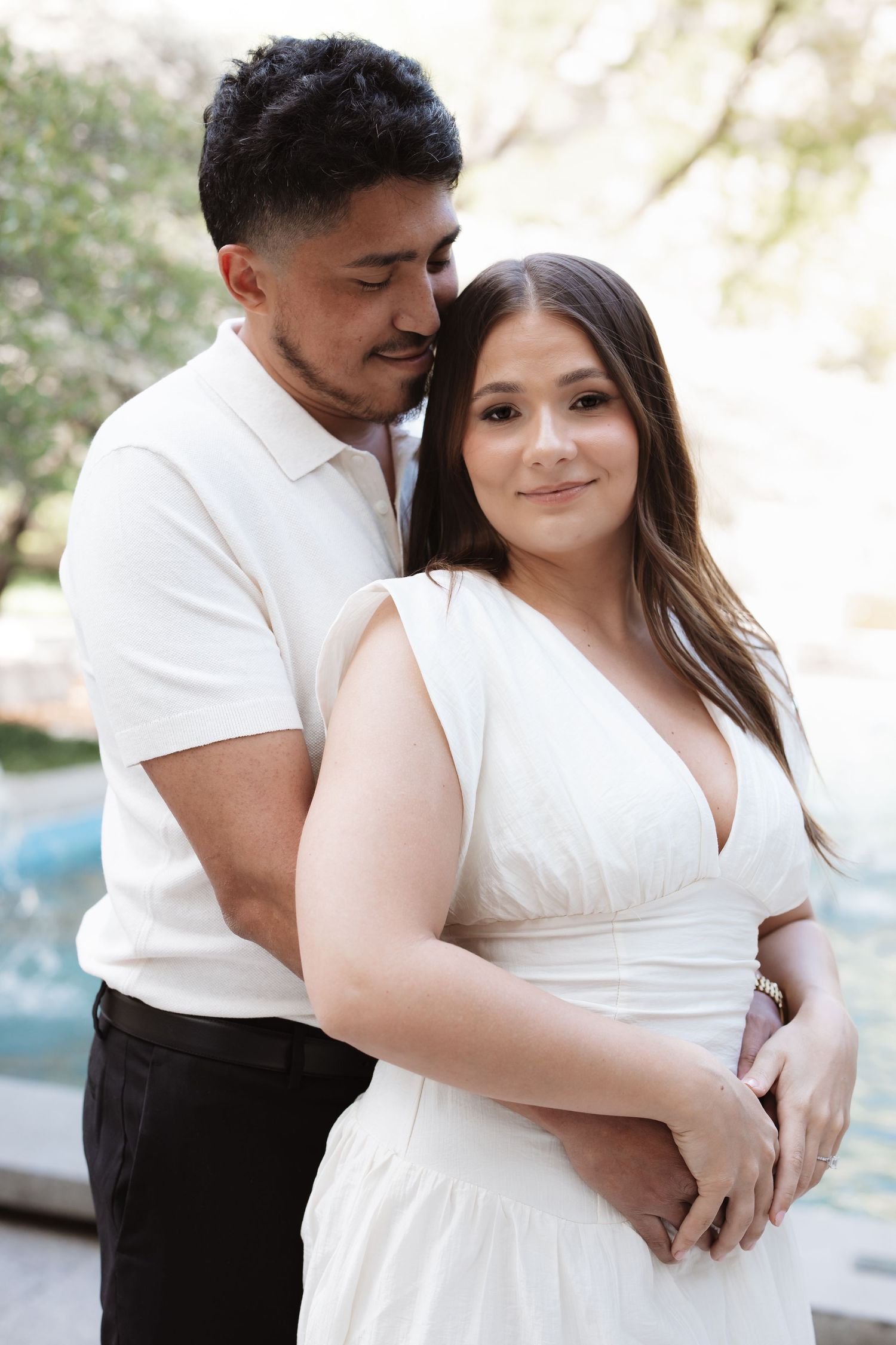 A couple in white outfits embracing outdoors, woman smiling at camera while man holds her close near a fountain.