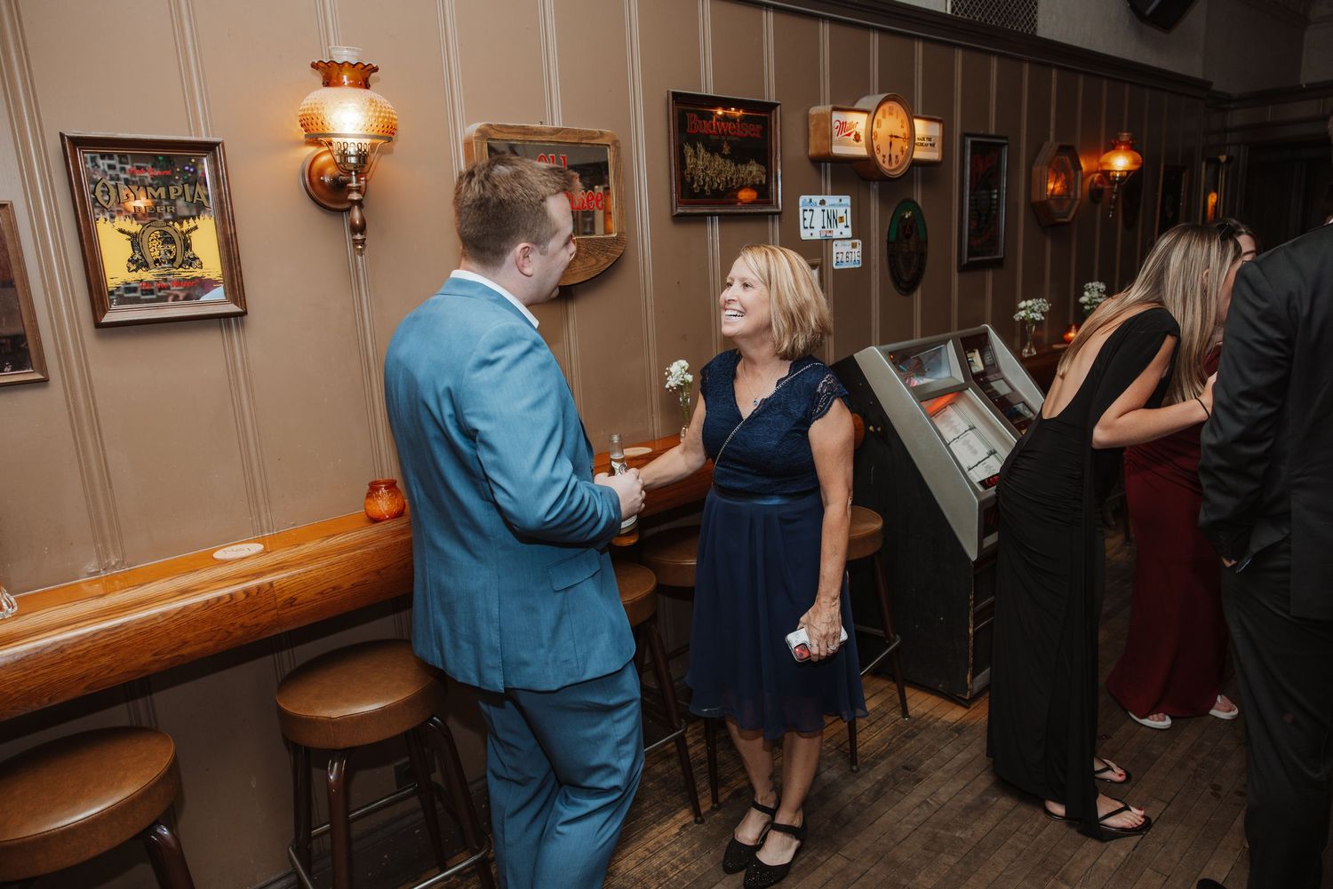Guests in formal attire socializing near a jukebox inside a warmly lit bar at a wedding reception.