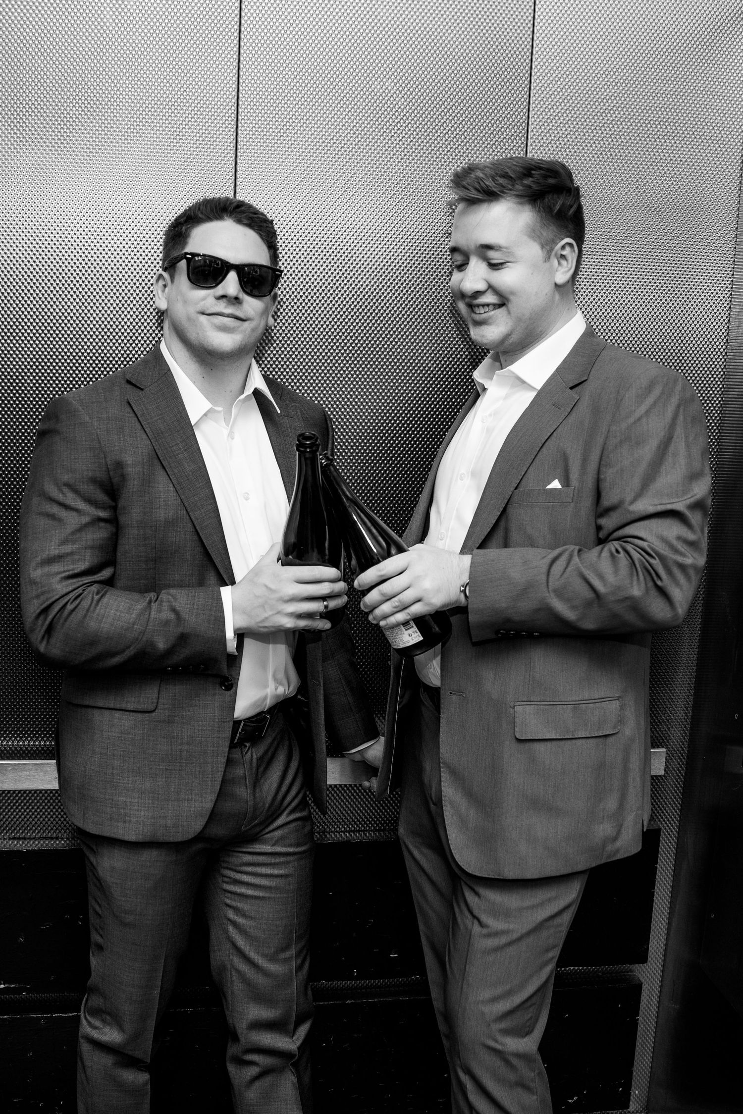 Two men in suits holding wine bottles smiling in black and white elevator photo.