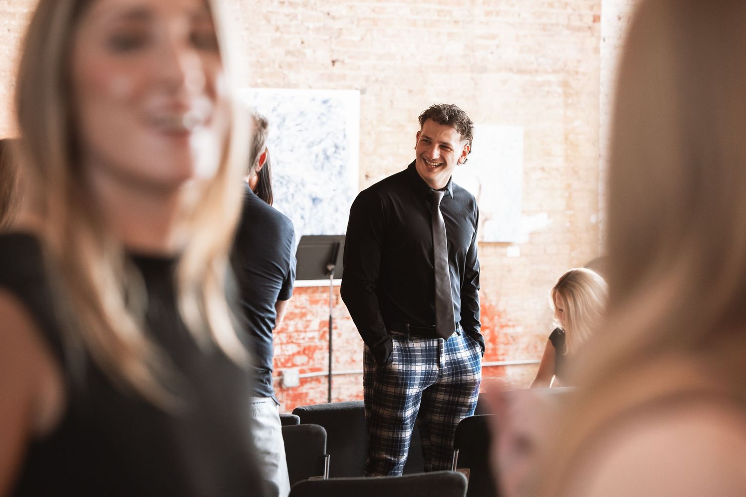 Man in black outfit smiling while standing among guests at an indoor social event.