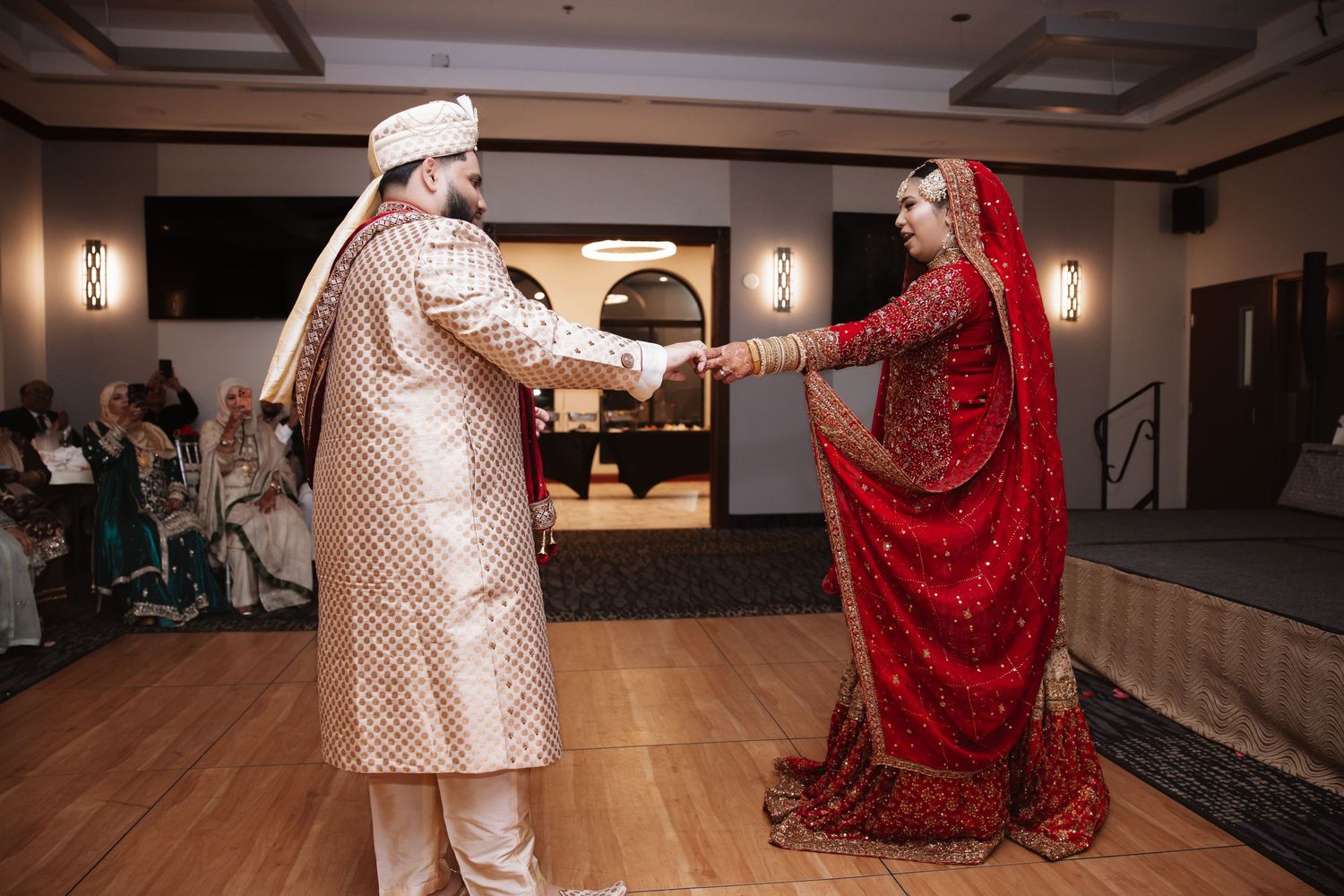South Asian bride in red lehenga and groom in gold sherwani holding hands during first dance.