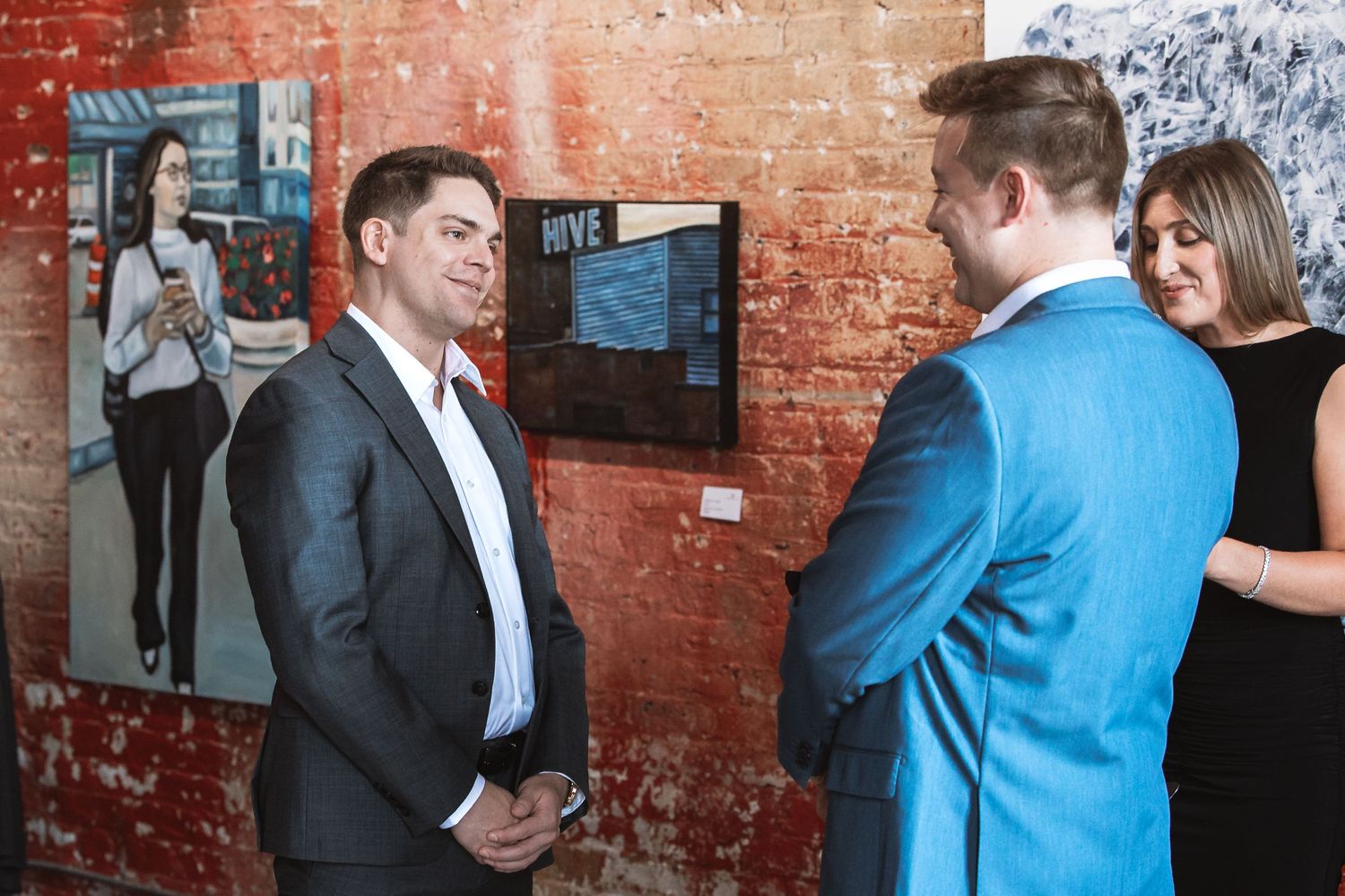 Two men in suits conversing at a wedding reception near brick wall artwork.
