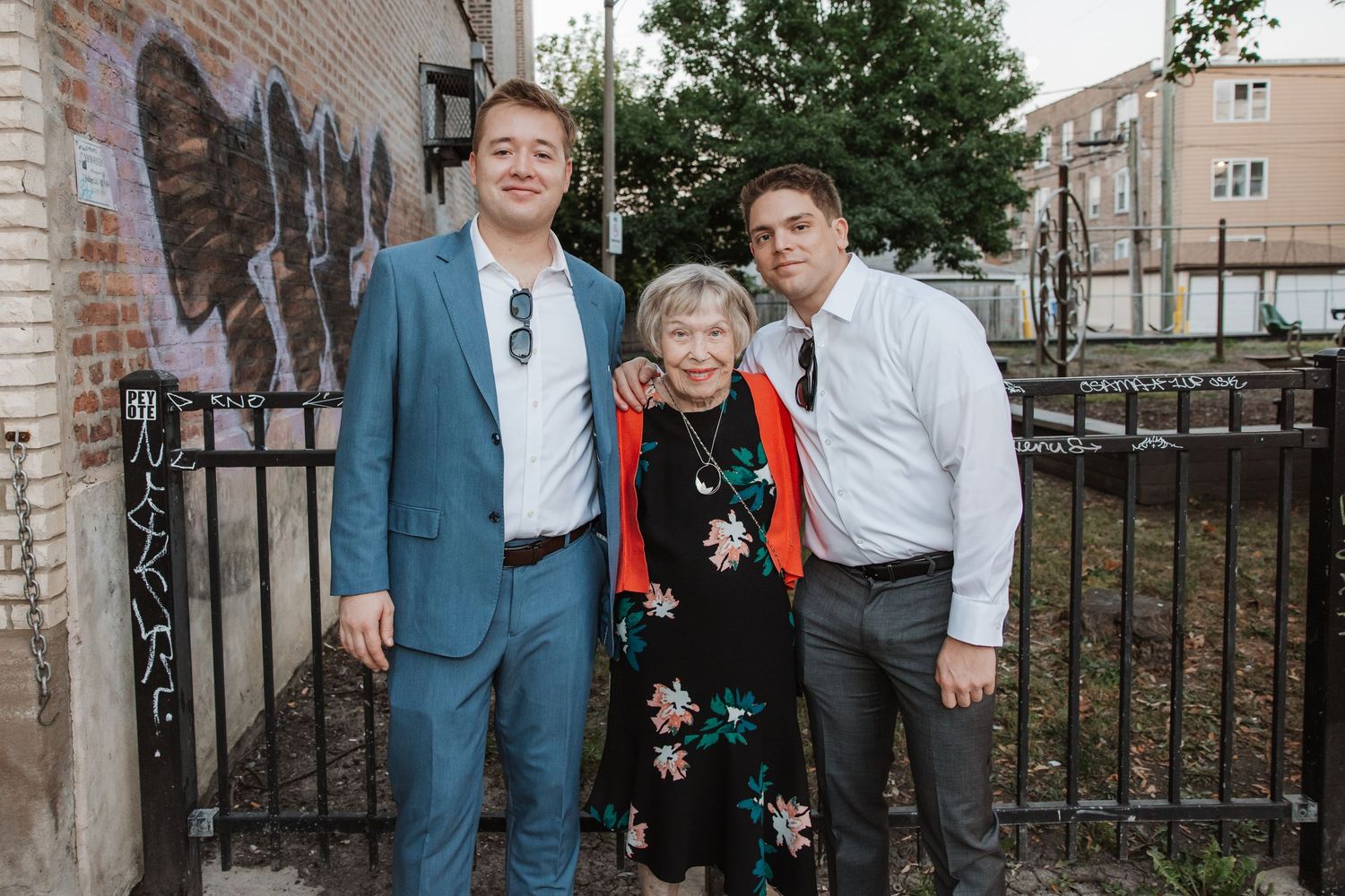 Two men in suits pose with elderly woman in floral dress outdoors near brick wall.