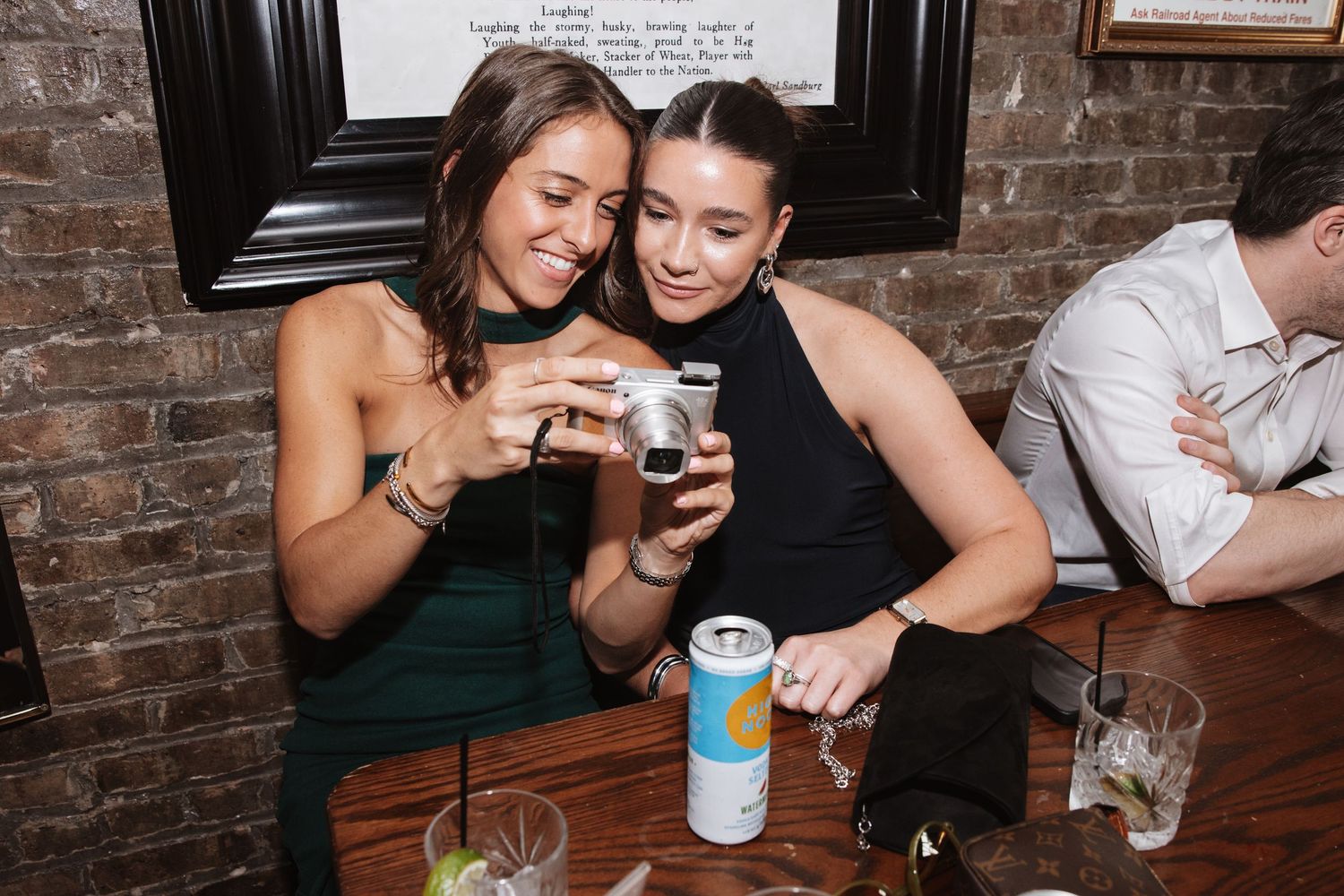Two young women looking at photos on a camera at a bar table.
