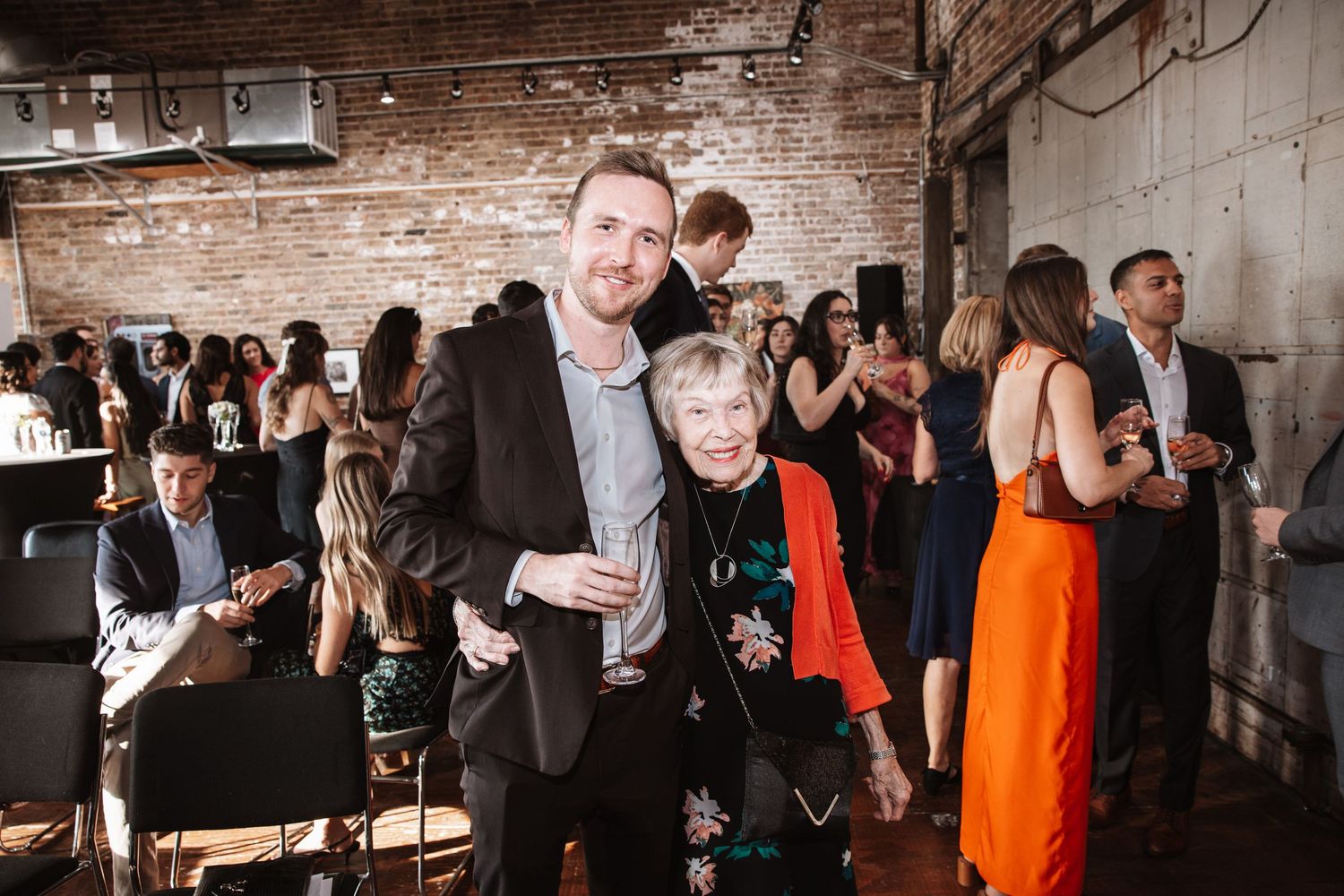 Man and elderly woman pose together smiling at a social gathering in a brick-walled venue.