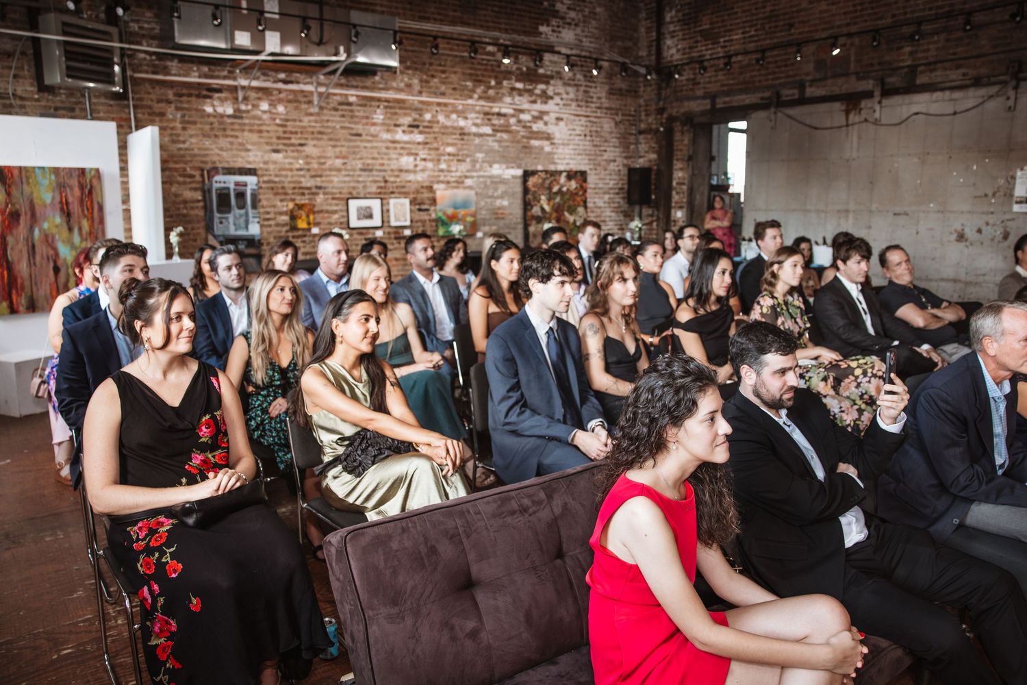 Large group of elegantly dressed wedding guests seated at indoor ceremony.