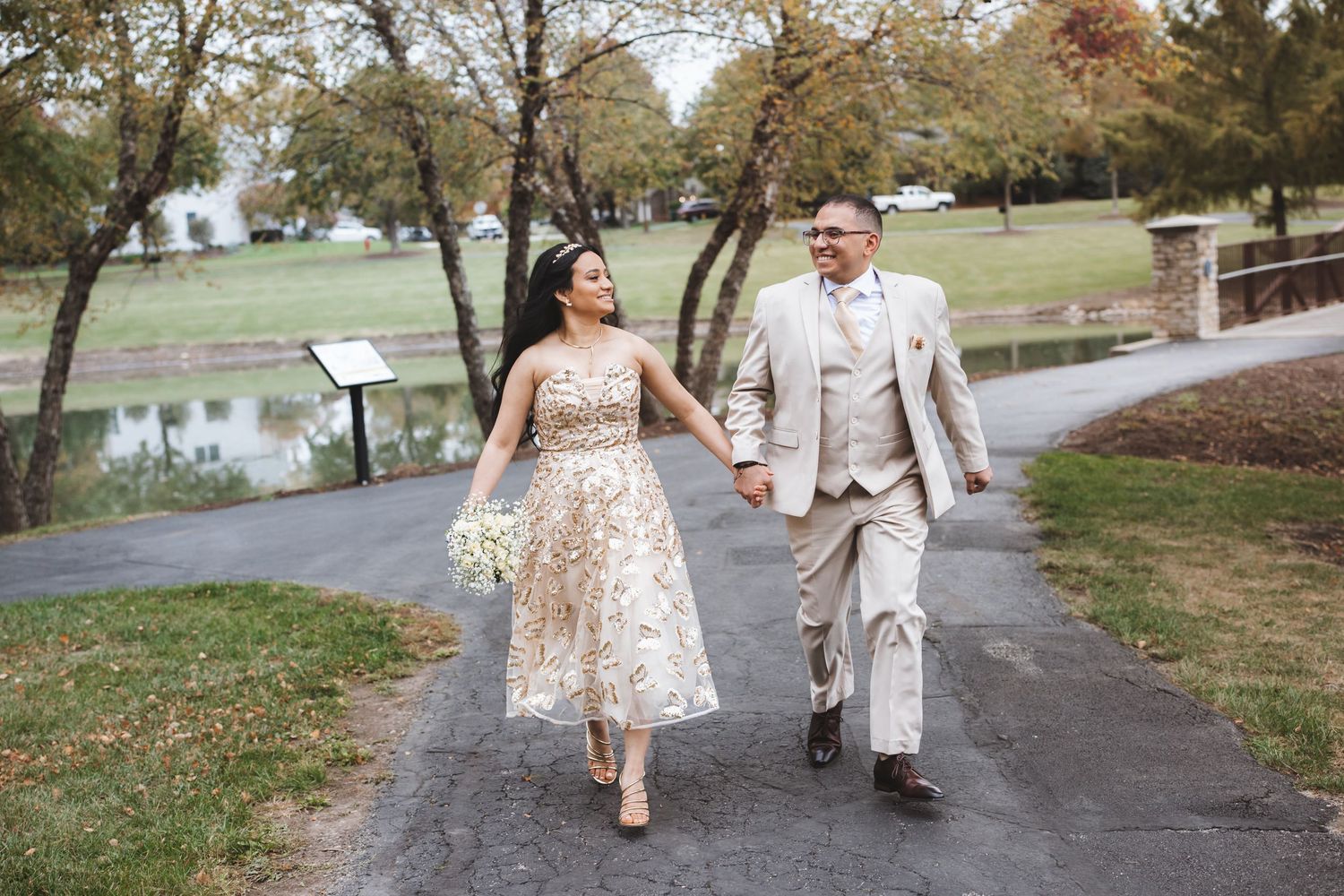 Happy newlyweds walking hand in hand on park path, bride holding white bouquet.