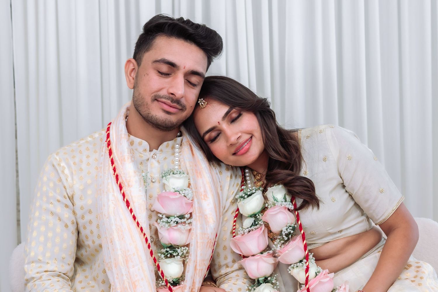 Indian bride and groom in white traditional wedding attire share a tender romantic moment wearing matching pink floral garlands.