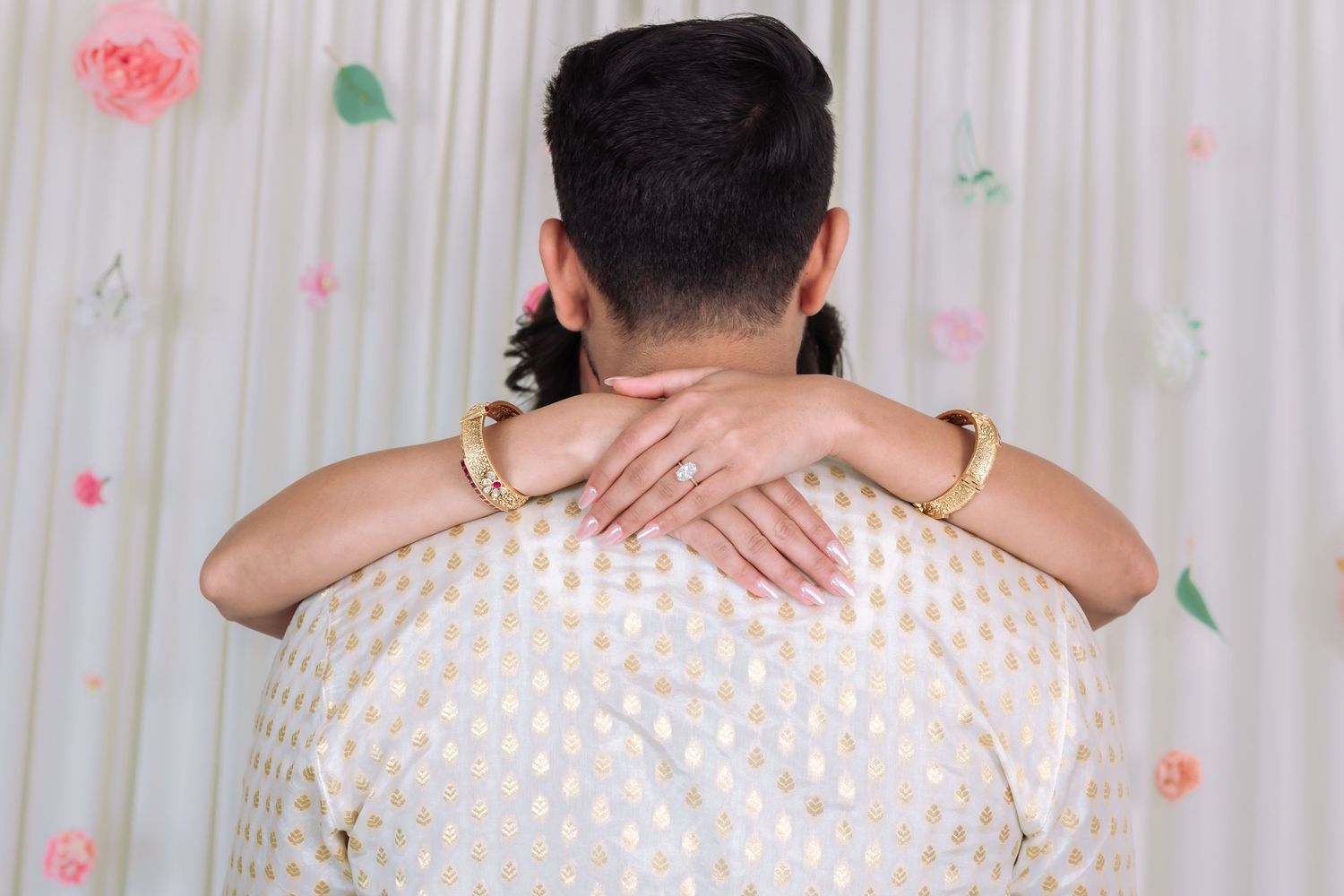 Couple embracing from behind at Indian engagement ceremony with floral backdrop.