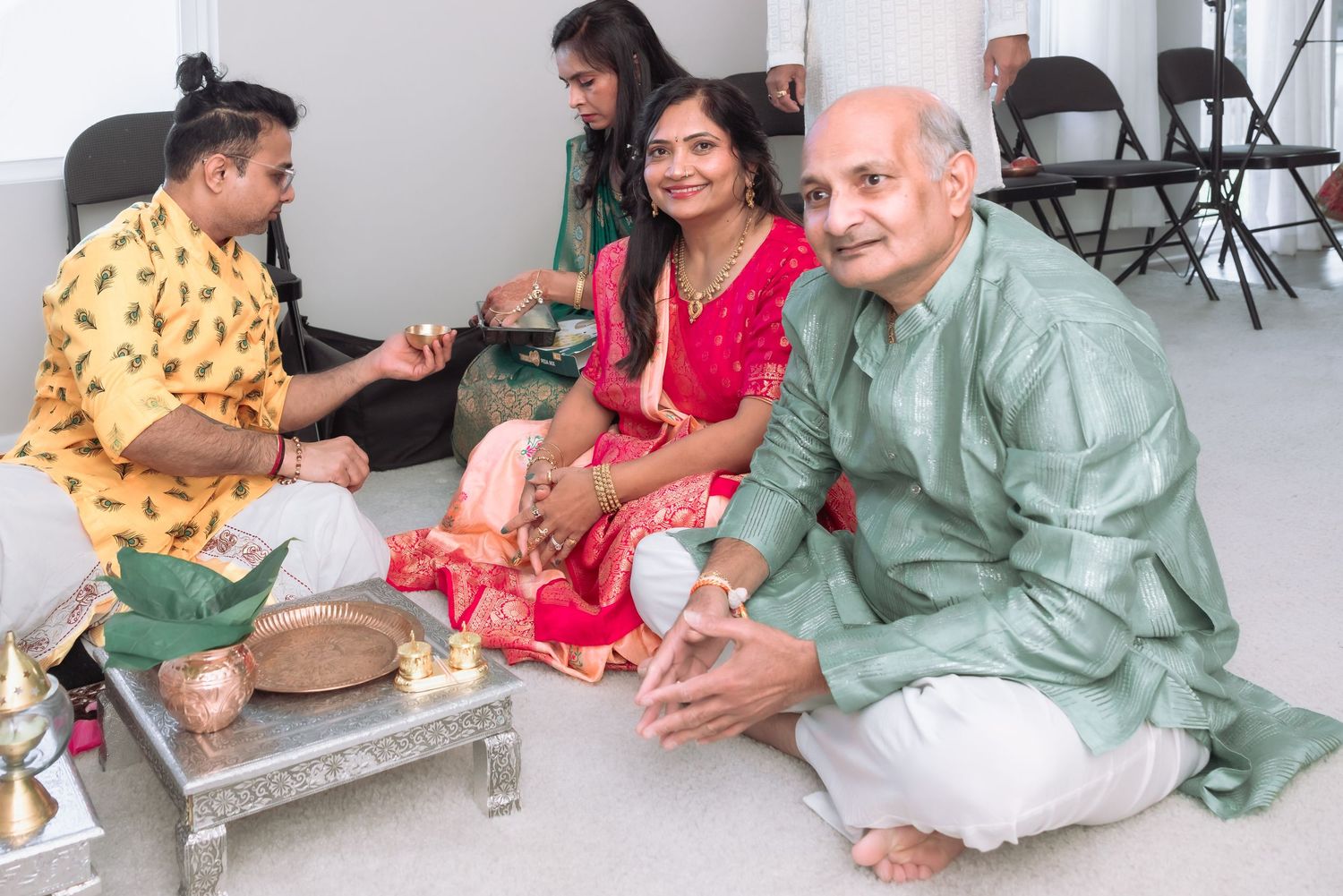 Indian family sitting on floor during traditional Hindu puja ceremony with ritual items on silver table.