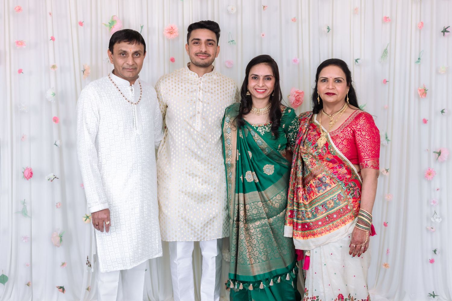 Indian family of four posing in traditional attire at wedding celebration with floral white backdrop.