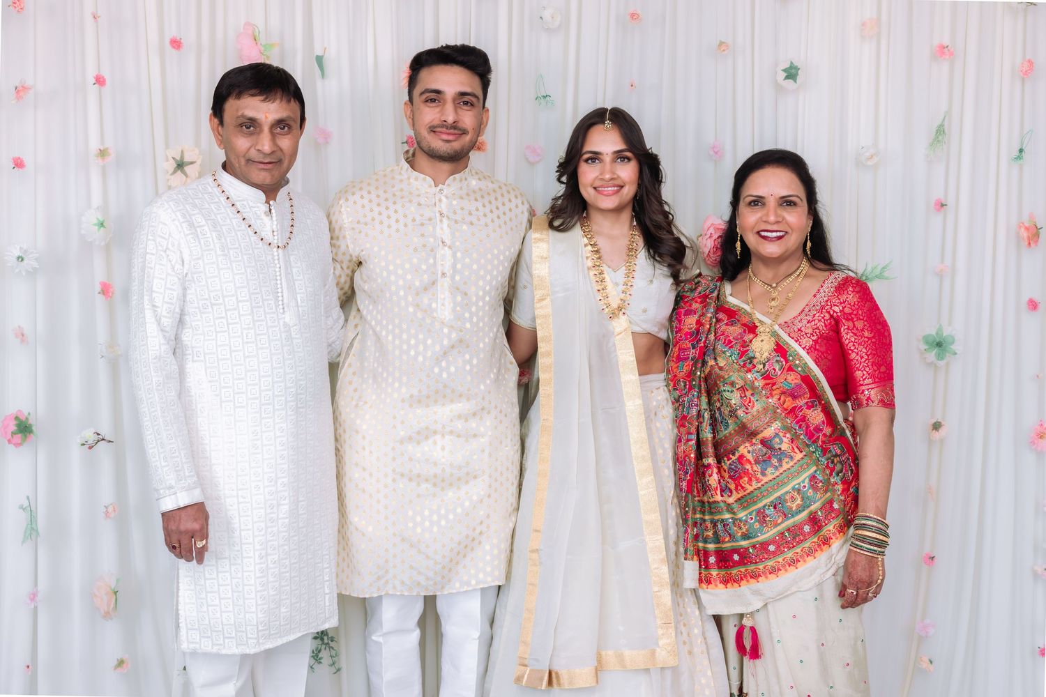 A family of four poses together in traditional Indian wedding attire against a floral backdrop.