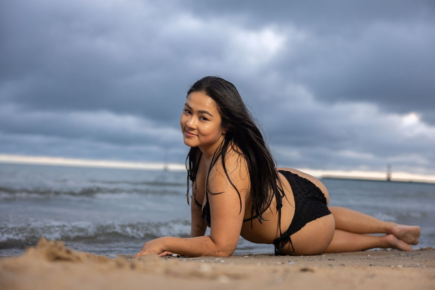 Young woman in black bikini lying on sandy beach smiling, with cloudy sky and ocean waves in background.