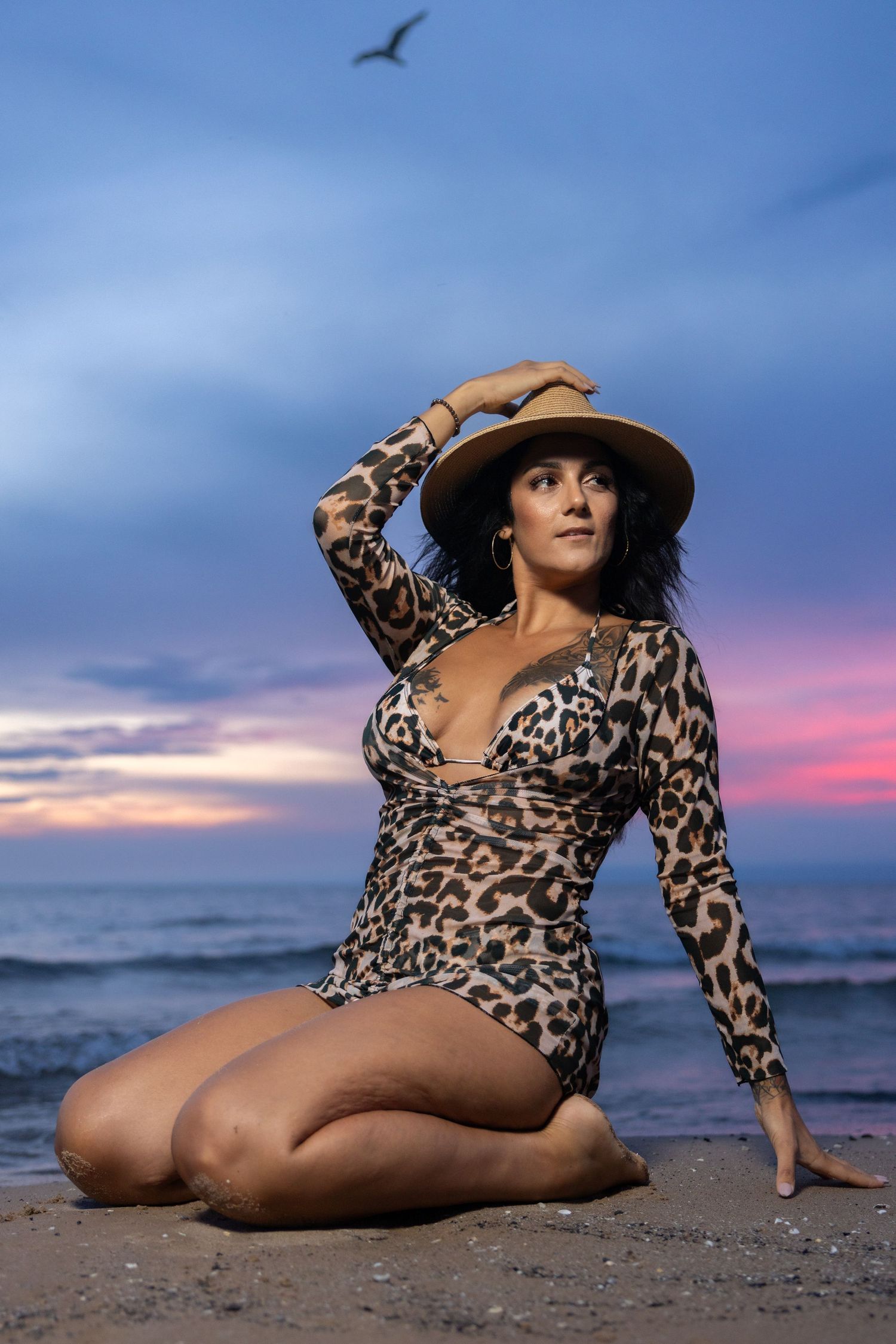 Woman in leopard swimsuit and hat kneeling on beach with bird in sky.