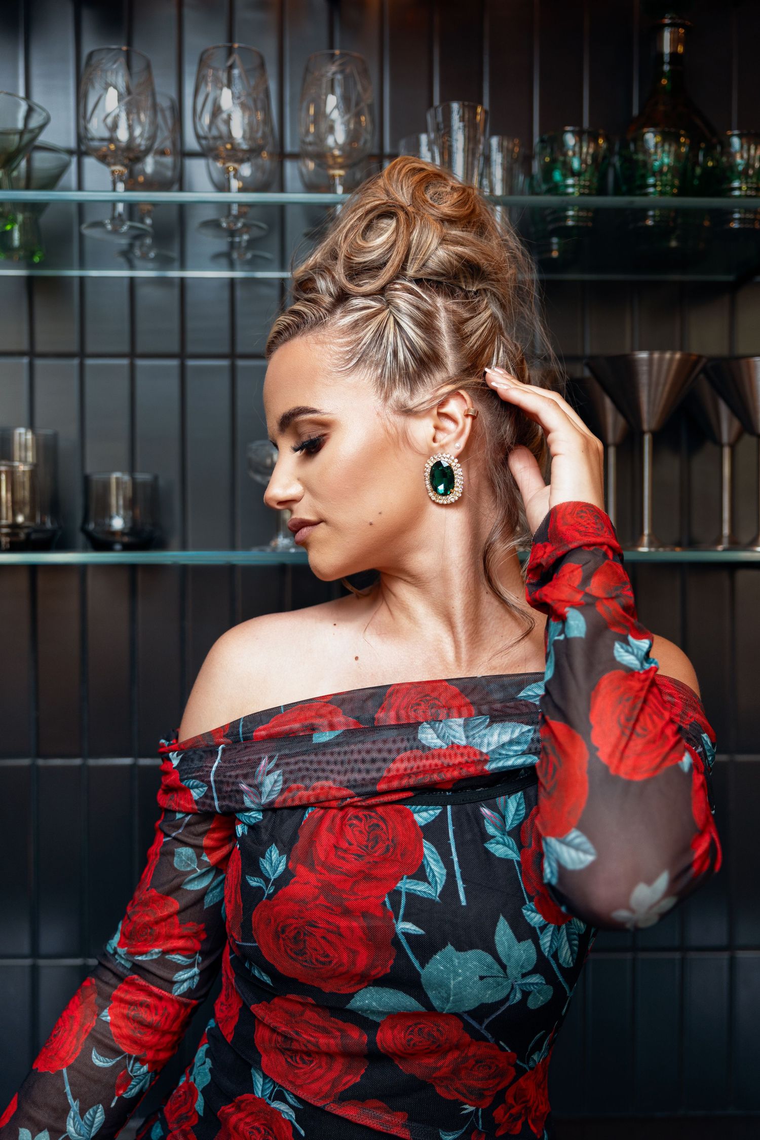 Blonde woman in off-shoulder rose print dress touching her updo hairstyle at a bar.