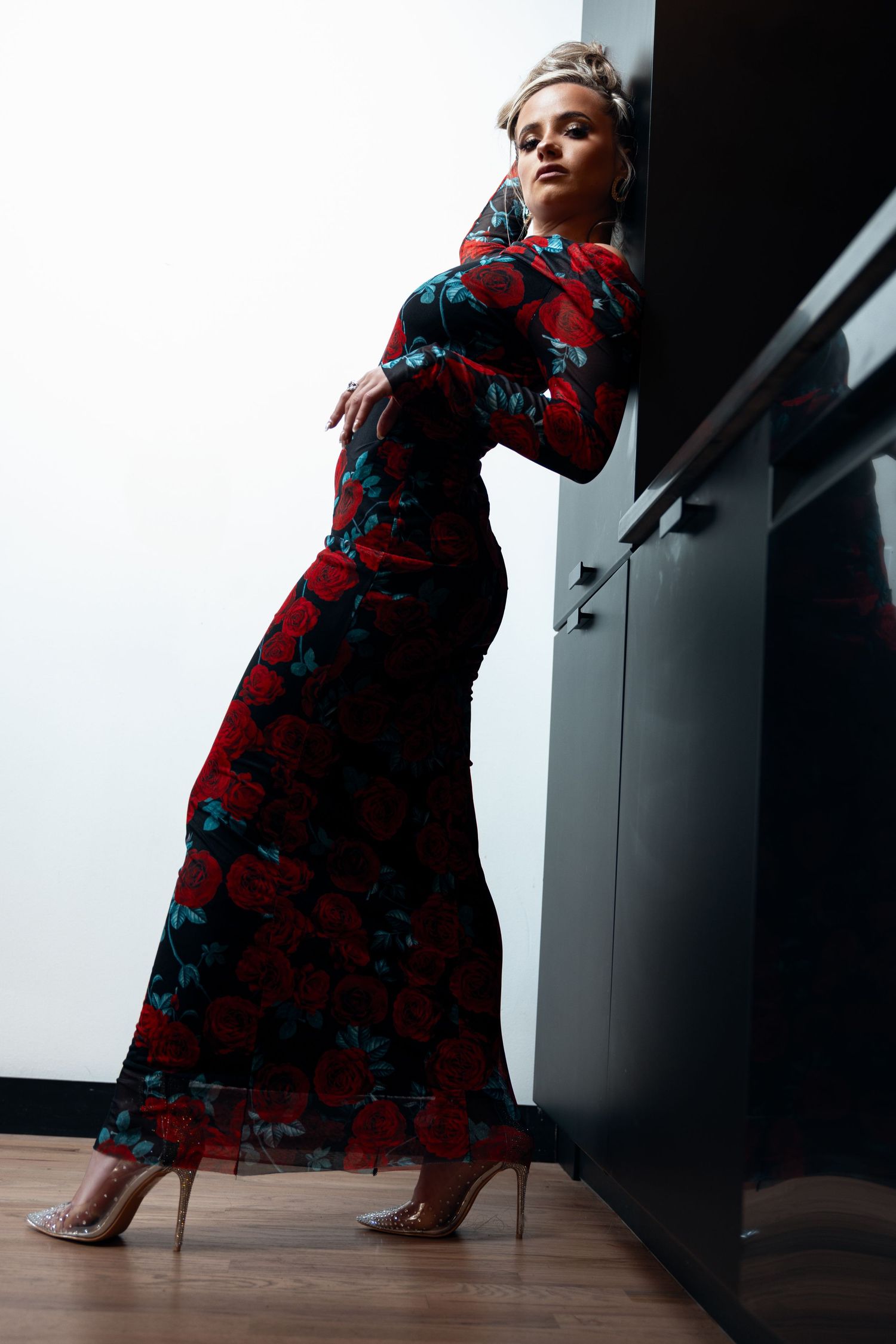 Blonde woman in red rose print floor-length dress leaning against dark cabinet in heels.
