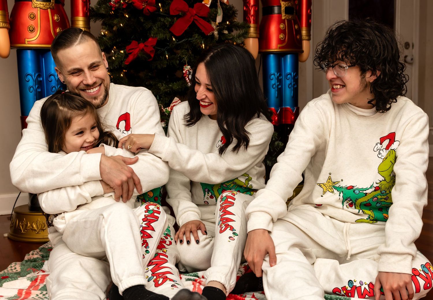 Happy family of four in matching Grinch Christmas pajamas sitting by decorated holiday tree.