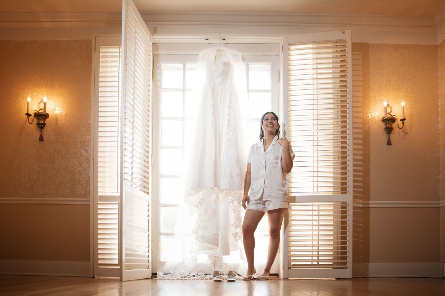 Bride in white robe standing beside her wedding gown hanging in a warmly lit room with shutters.