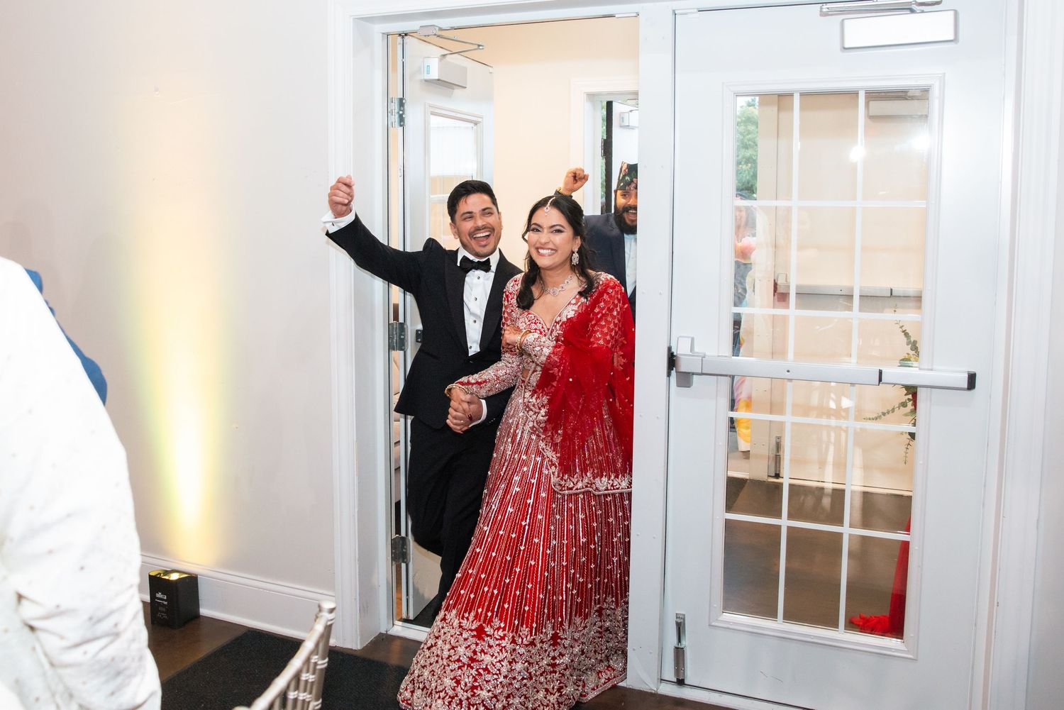 Bride in red lehenga and groom in tuxedo make grand entrance at Indian wedding reception.