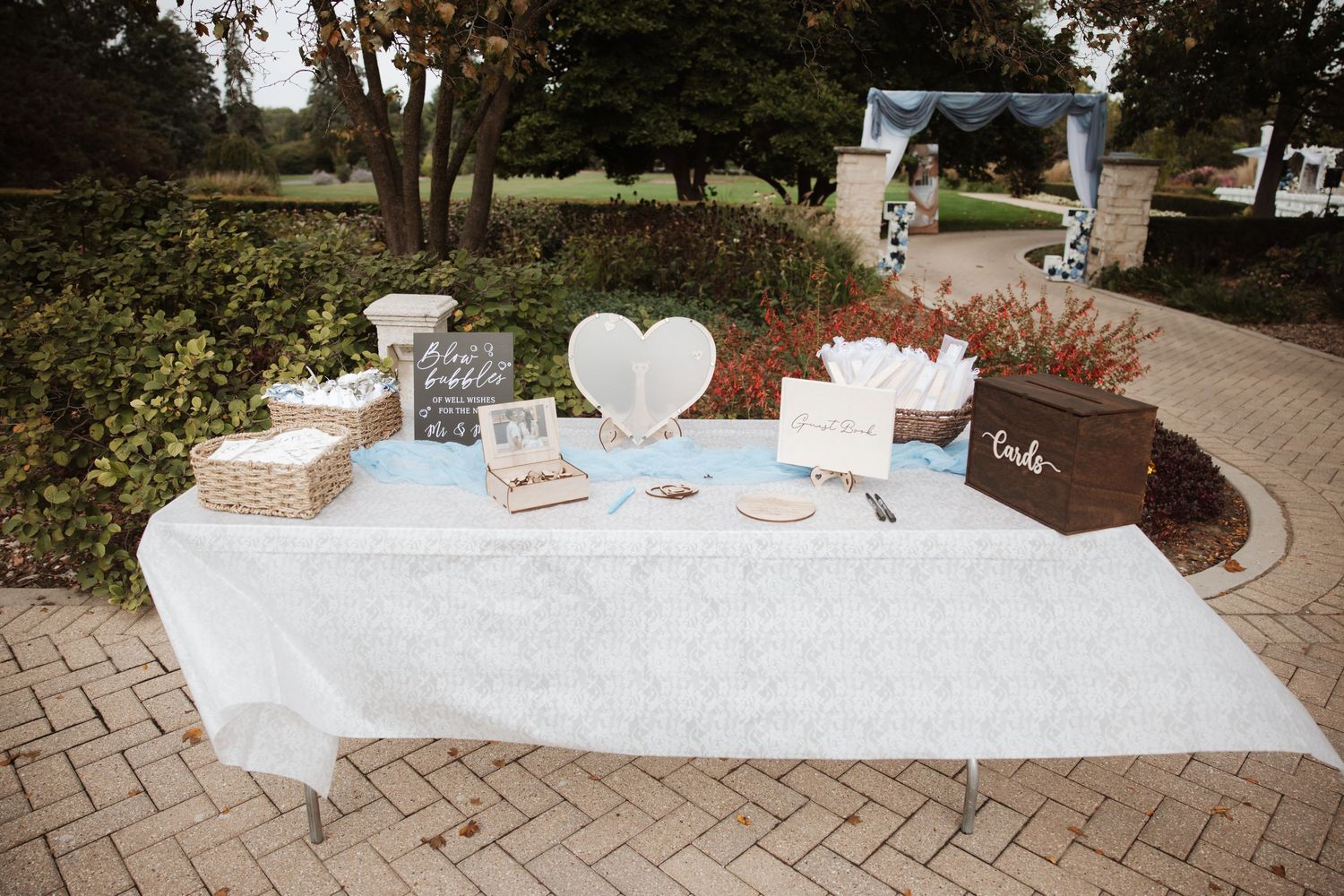 Outdoor wedding reception table with heart decor, guest book, and floral arch in background.