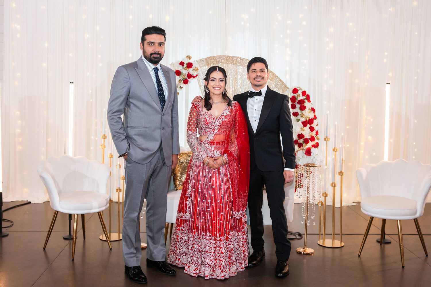 Three people posing at elegant Indian wedding reception with red floral backdrop and white decor.