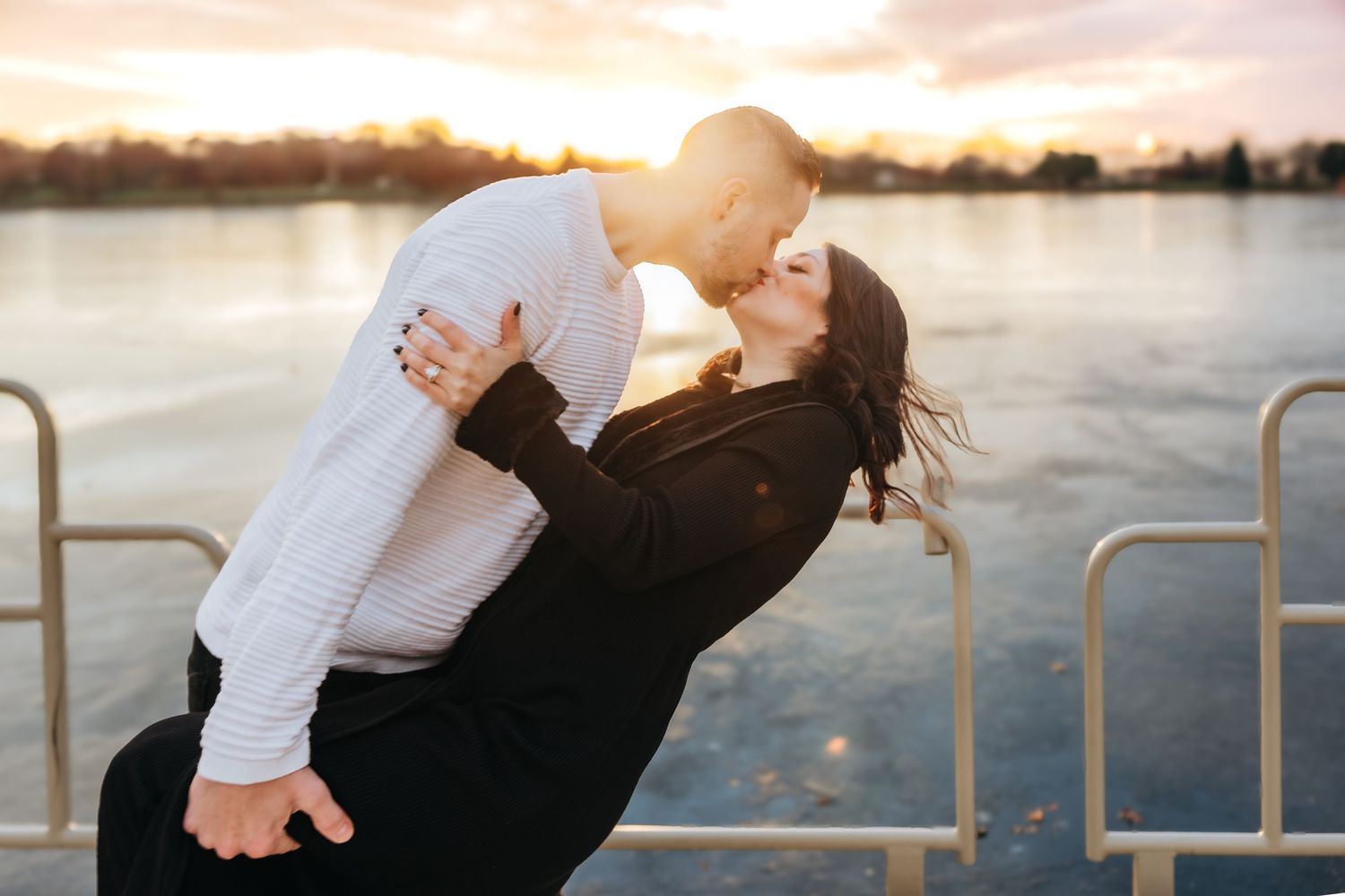 Couple sharing a romantic dip kiss at sunset by a serene waterfront, woman in black dress and man in white sweater.