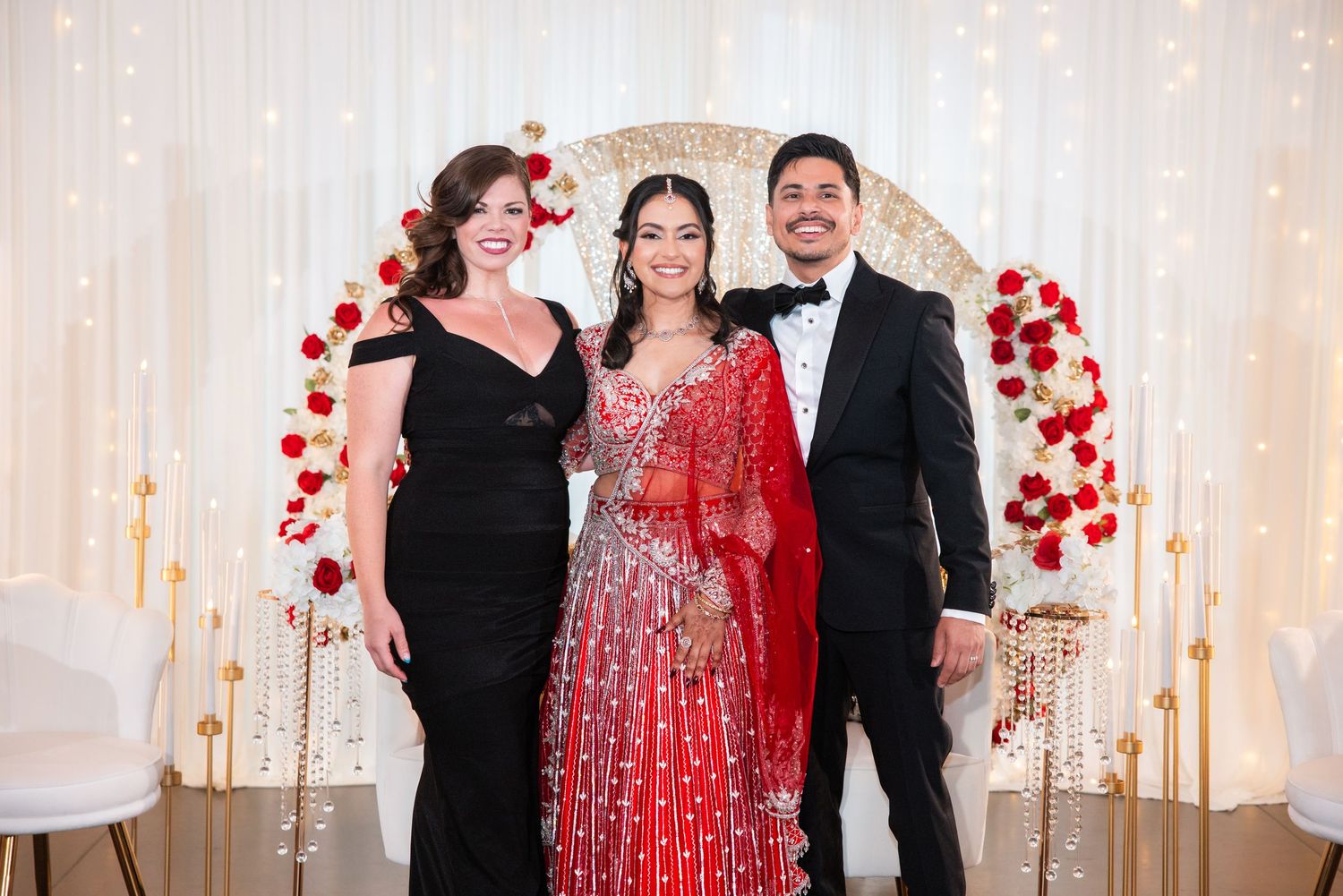 Bride in red Indian bridal lehenga posing with two guests at decorated wedding backdrop.