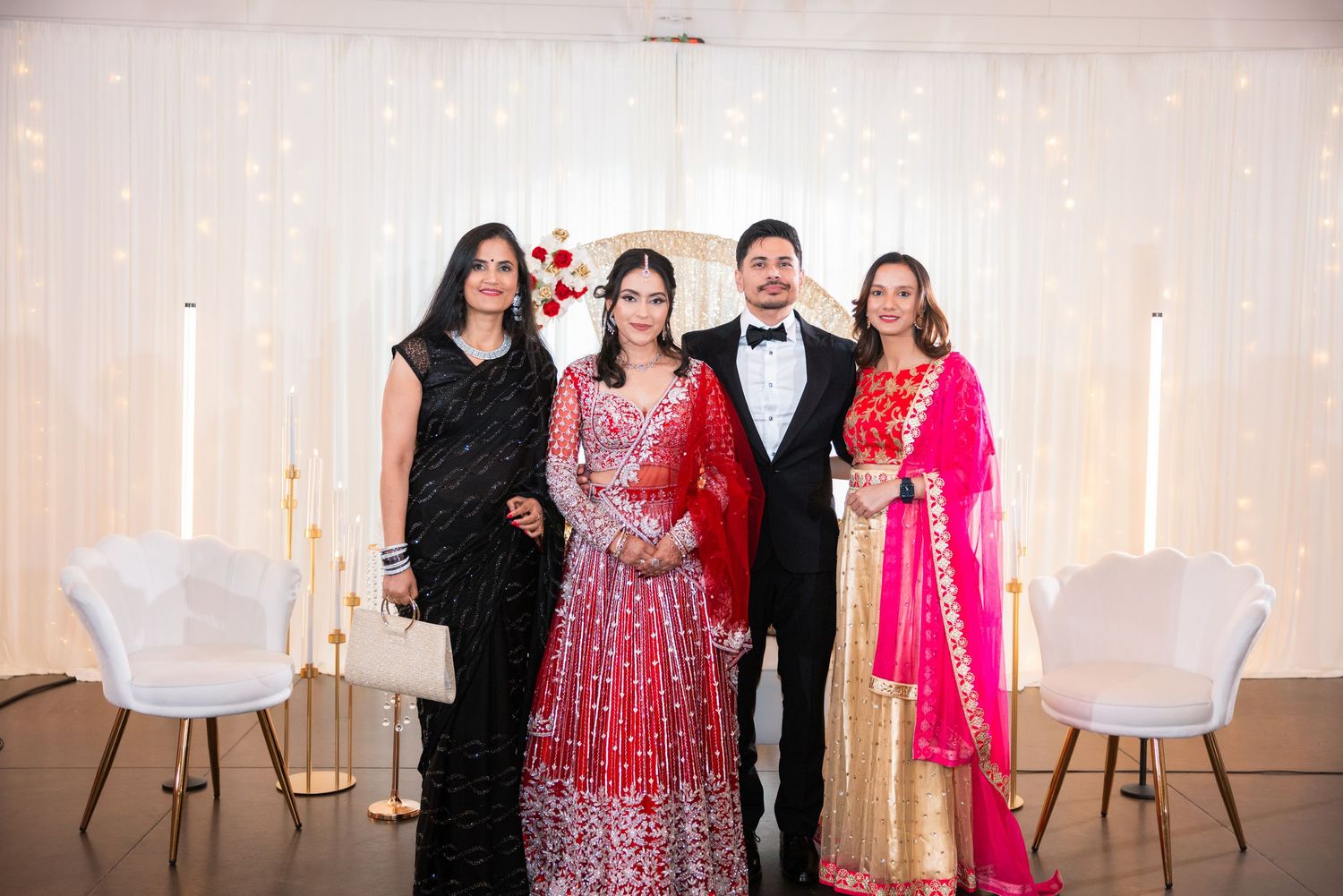 Indian bride in red lehenga with groom in tuxedo and two women guests at wedding.