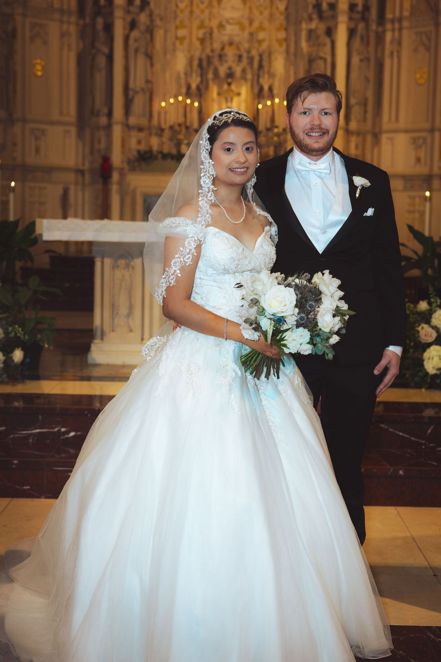 Smiling bride and groom posing together in ornate church interior, bride holding white floral bouquet.