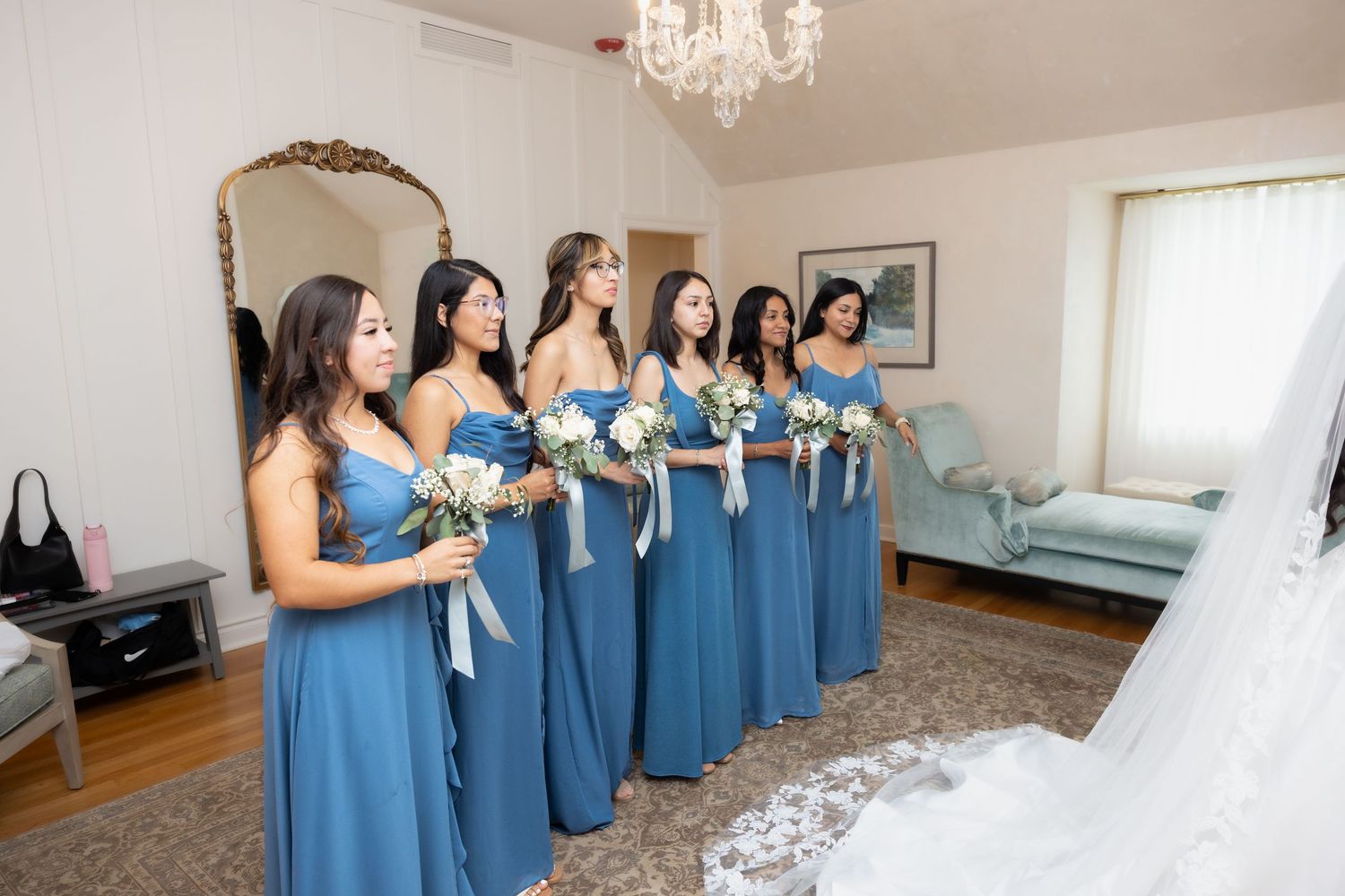 Six bridesmaids in dusty blue gowns holding white bouquets react to seeing bride before wedding ceremony.