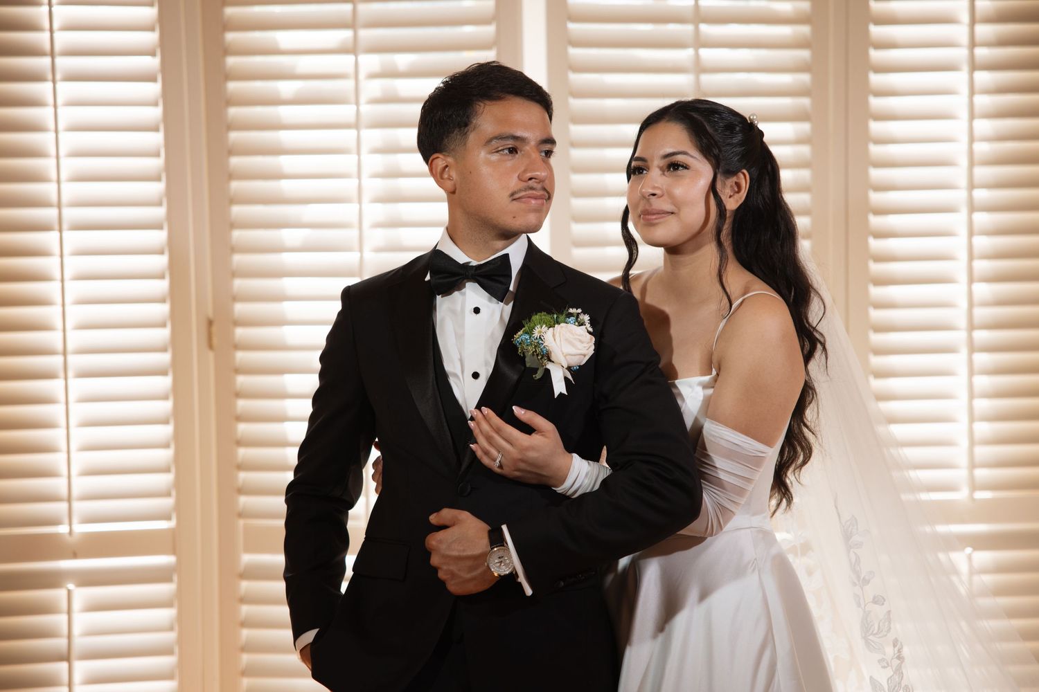 Bride in strapless gown holding groom's arm as they pose together in front of wooden shuttered blinds.
