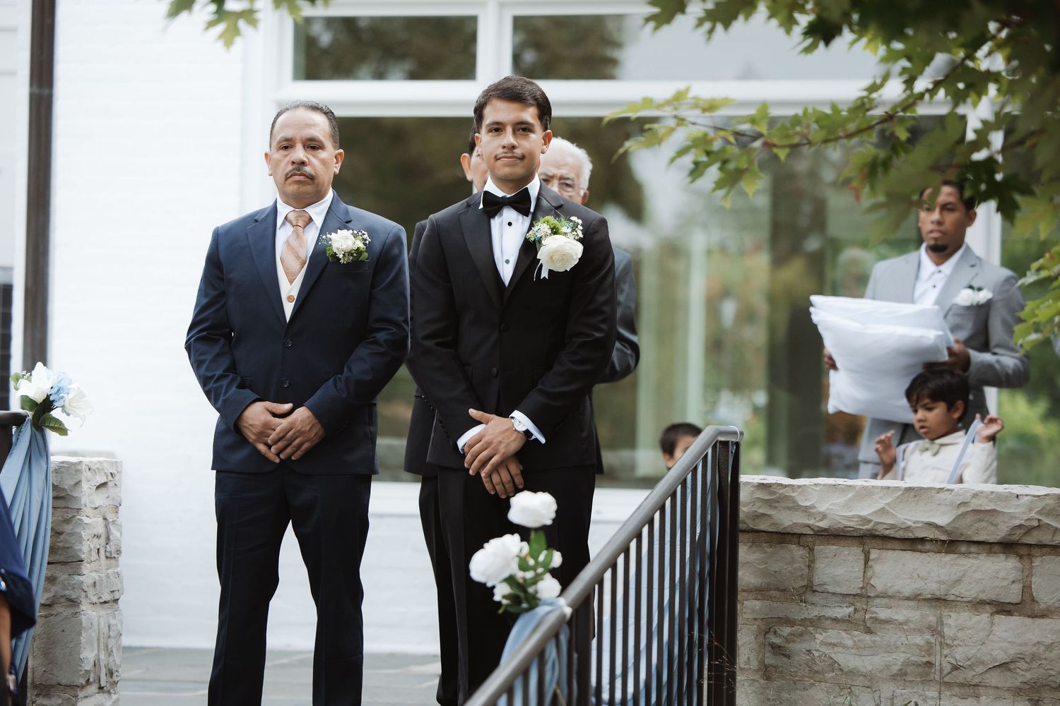 Groom in black tuxedo stands with father wearing boutonniere at outdoor wedding ceremony.