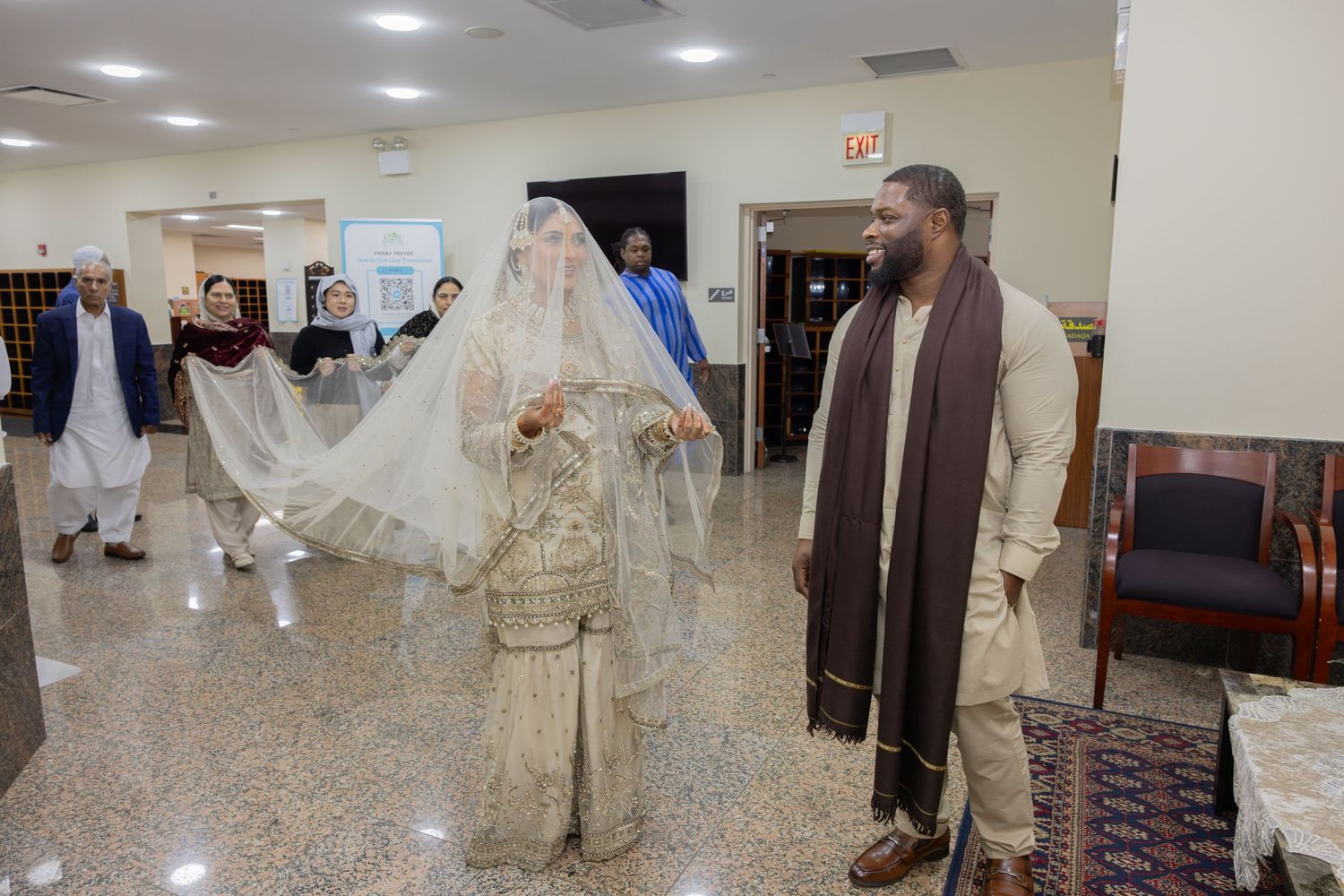 A South Asian bride in white embroidered attire walks beside her groom in a brown shawl indoors.