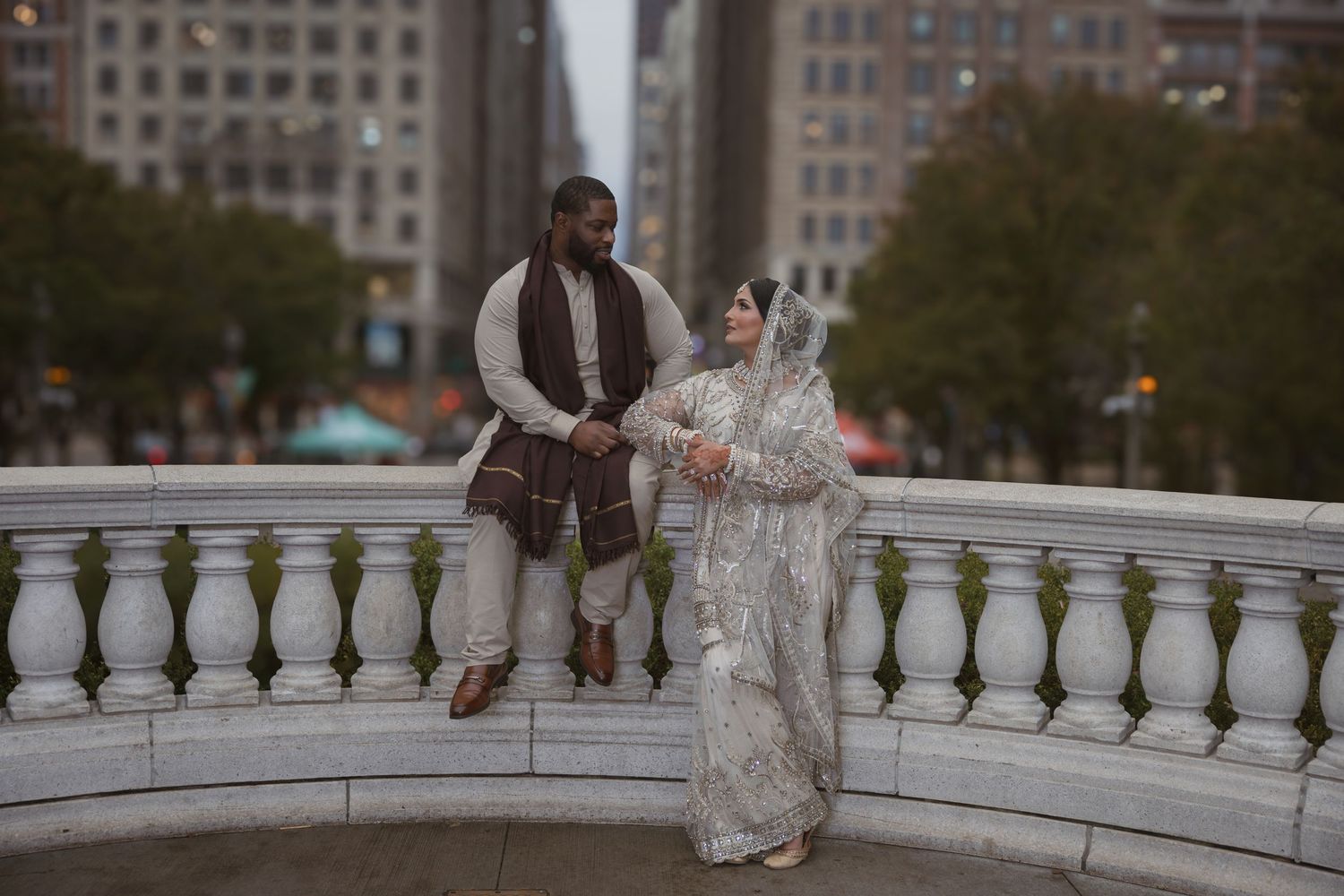 Newlyweds in traditional South Asian wedding attire share romantic moment on city bridge.