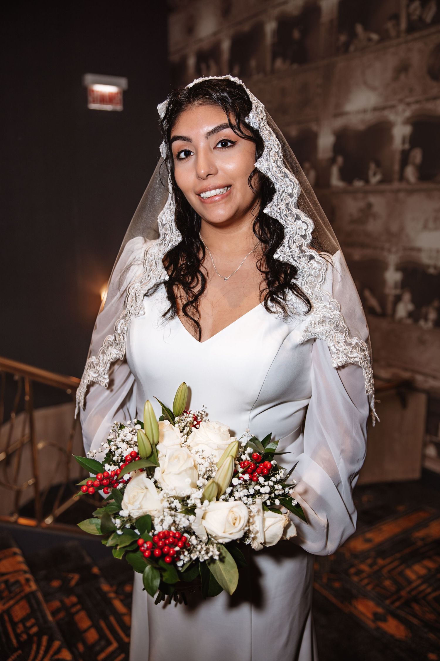 Radiant bride in white gown and lace veil holds bouquet of white roses and red berries in dark venue.