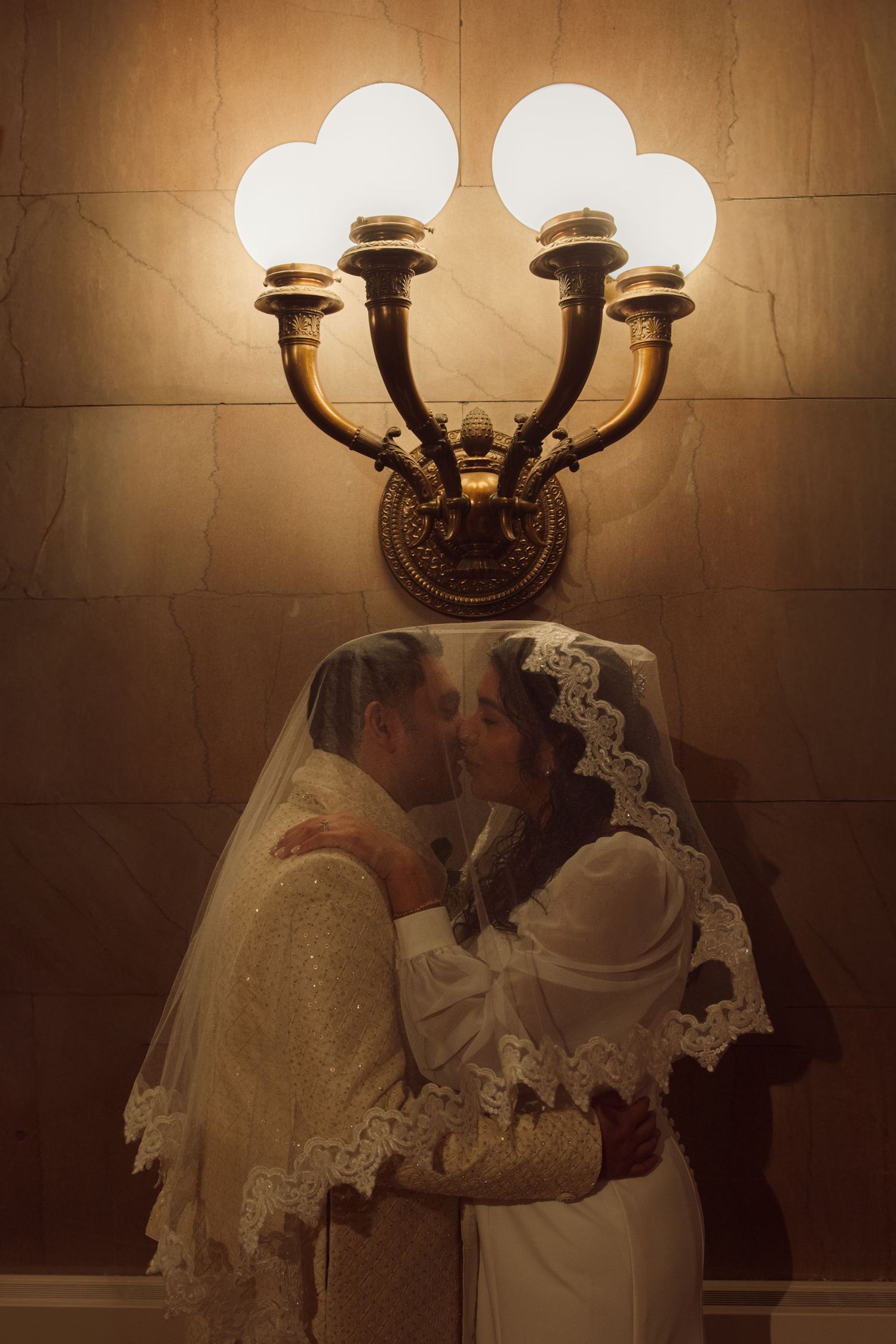 Bride and groom sharing a romantic kiss under a glowing brass wall sconce, both wearing white wedding attire with lace veils.