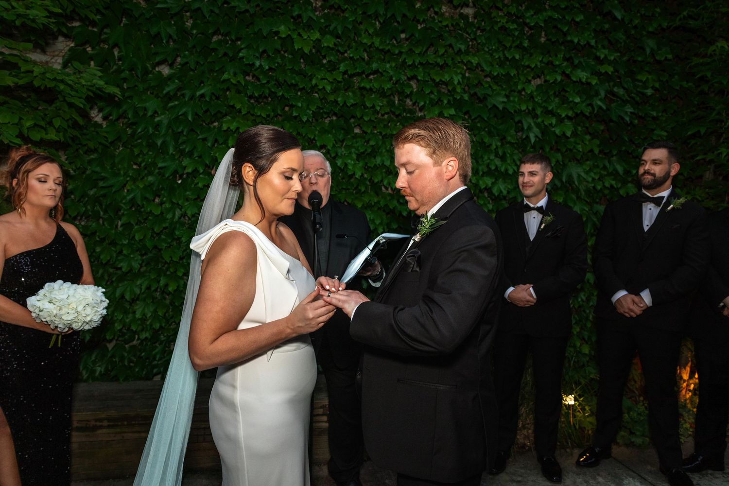 Groom places wedding ring on brides finger during outdoor ceremony with wedding party.