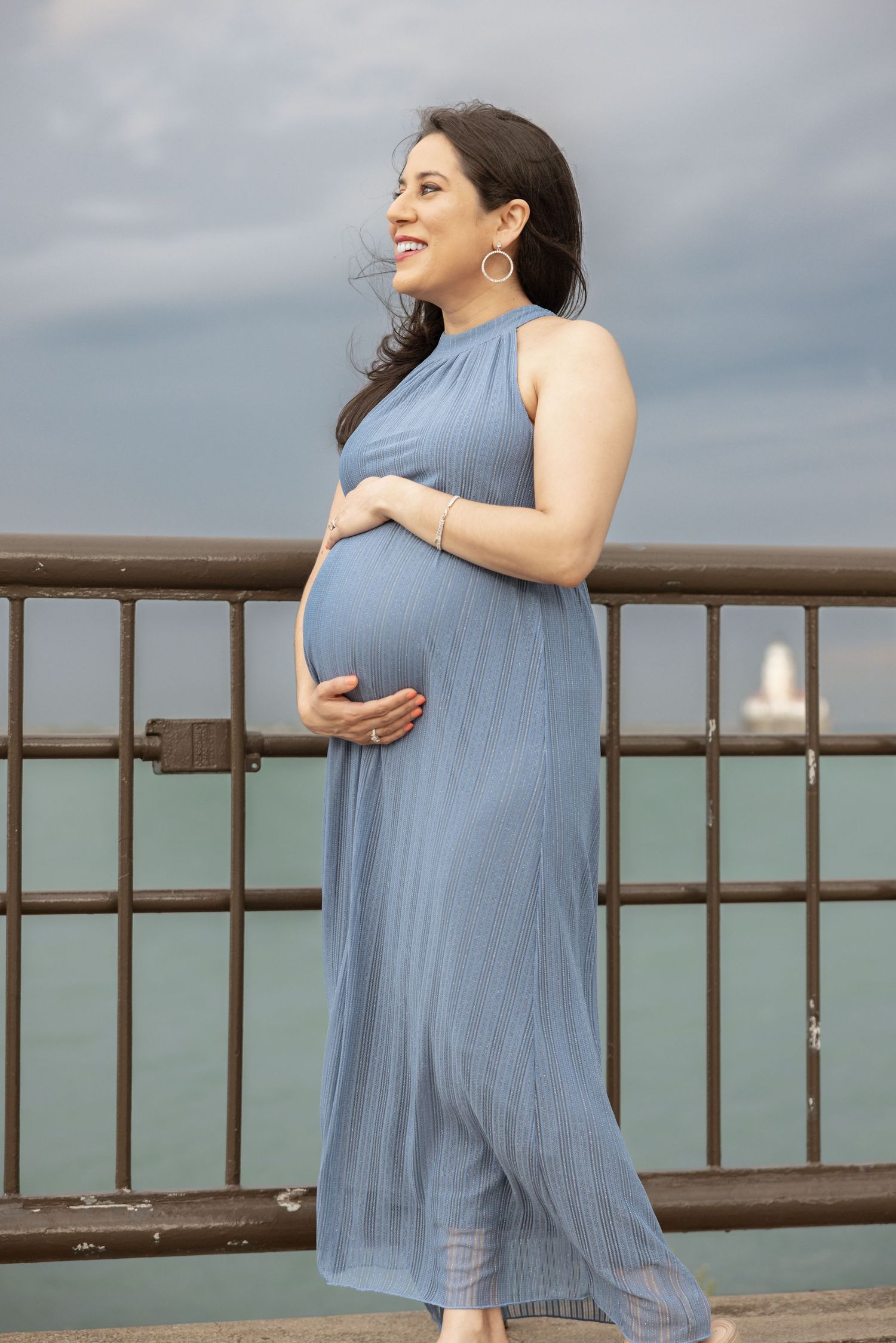 Pregnant woman in blue maxi dress cradling belly on pier with ocean view.