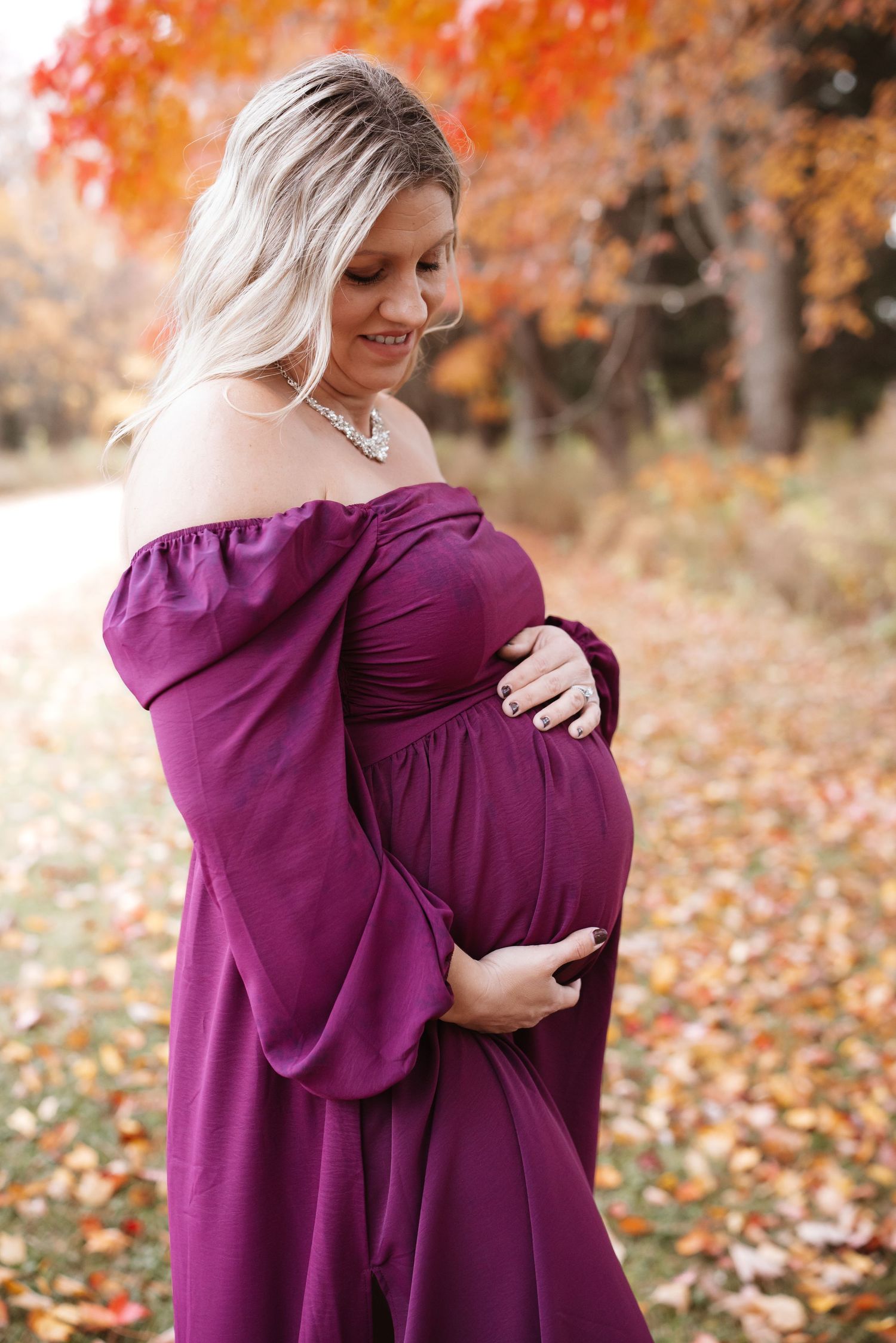 Pregnant woman in purple off-shoulder dress gently holding baby bump amid colorful fall foliage.