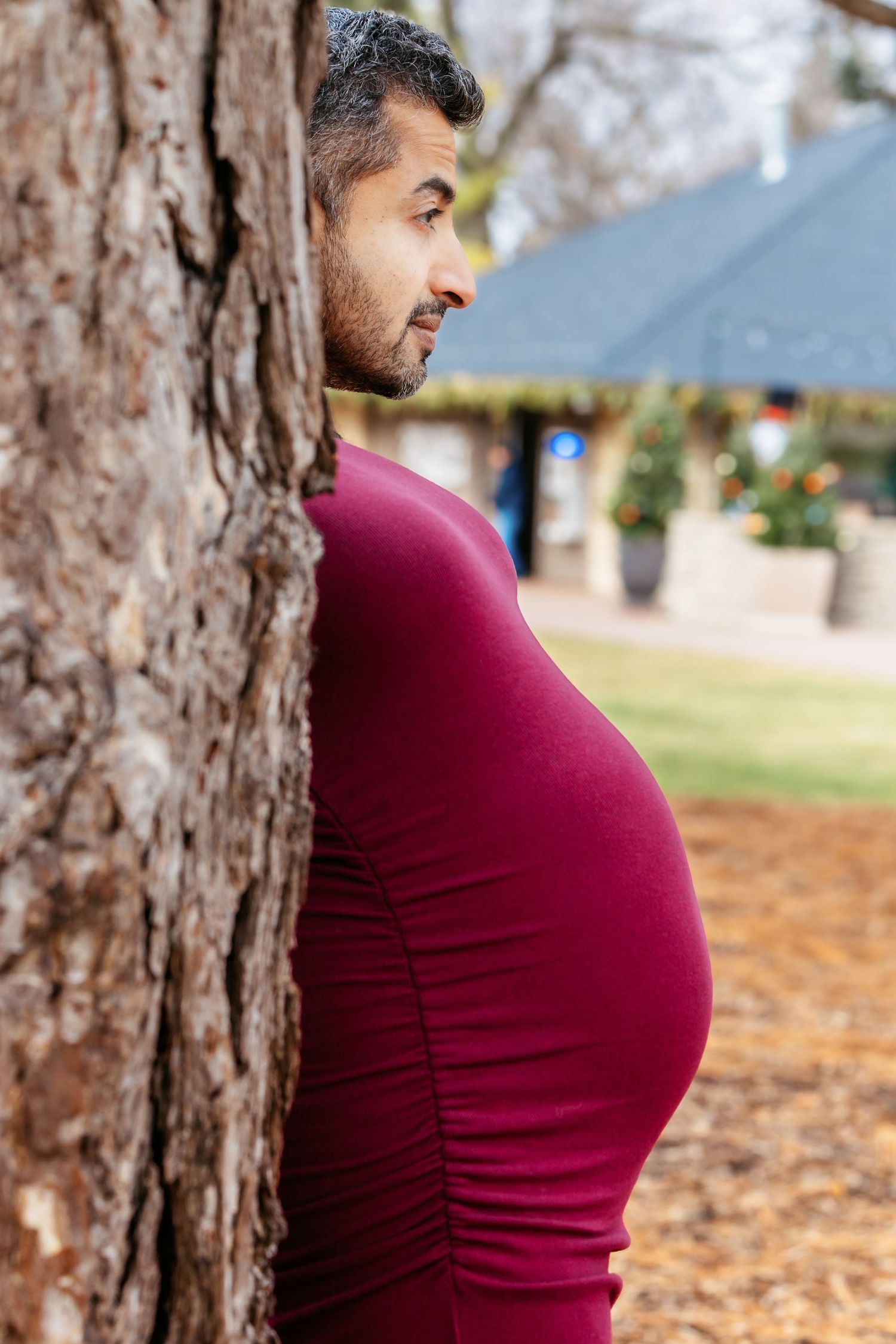 Side view of pregnant woman in red dress standing beside a large tree in a park.